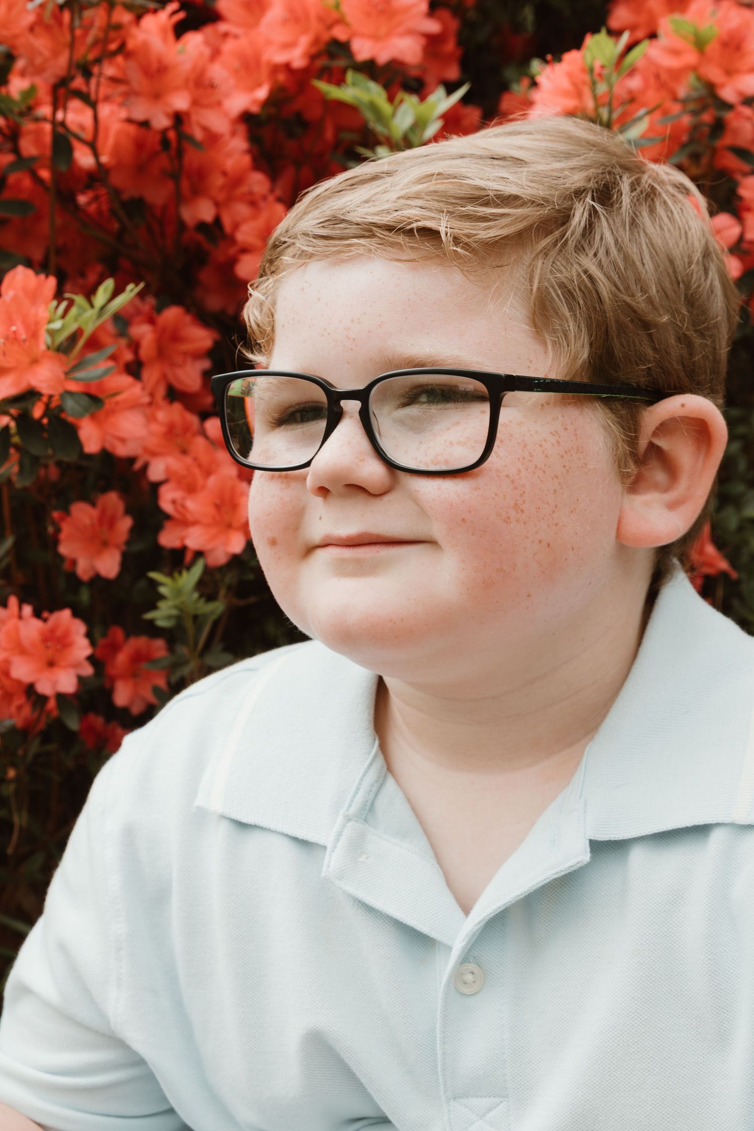 A close-up portrait showing a gentle smile against a backdrop of bright coral-colored flowers.