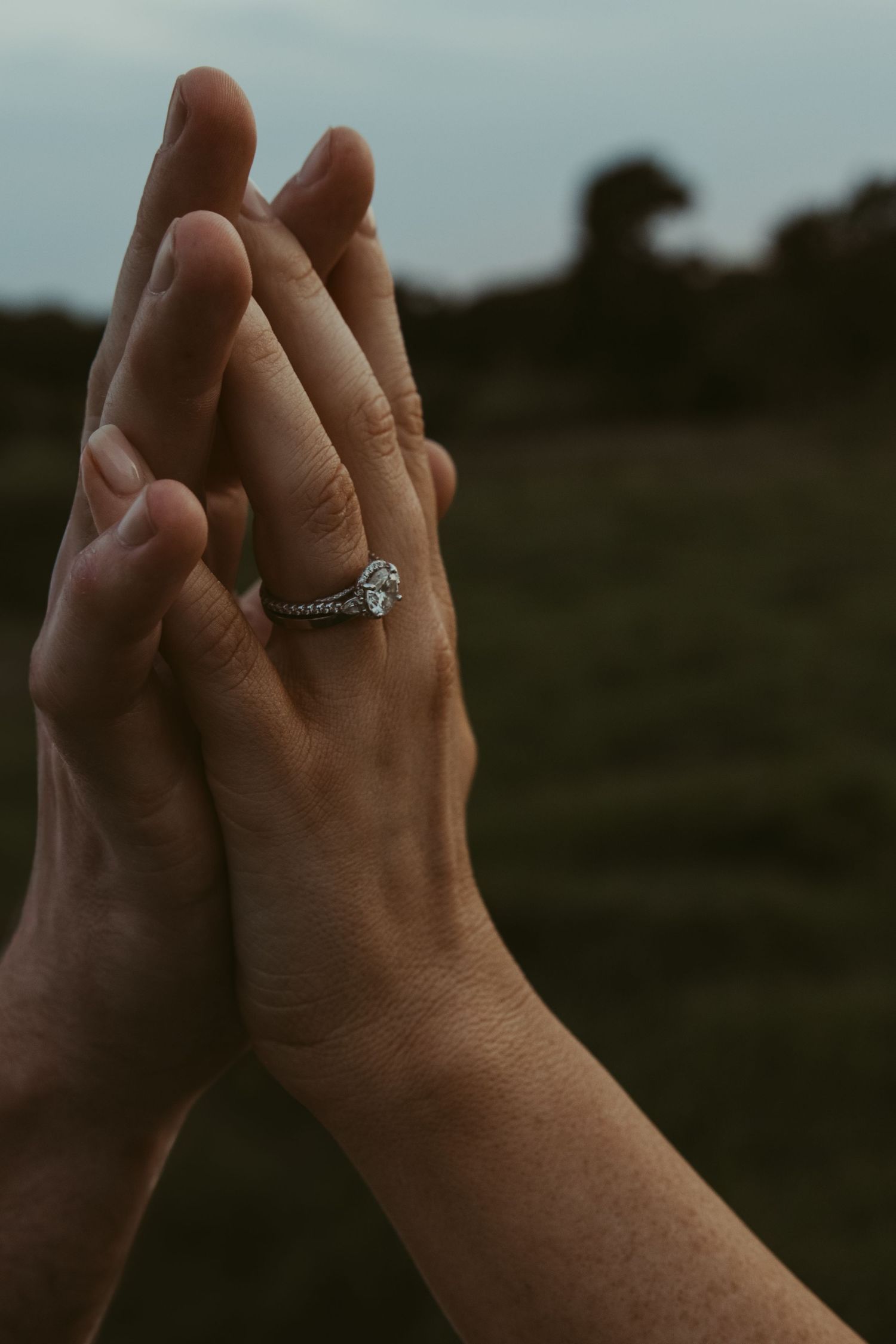 Two hands clasped together against a blurry dark background, with one hand wearing a sparkly engagement ring.