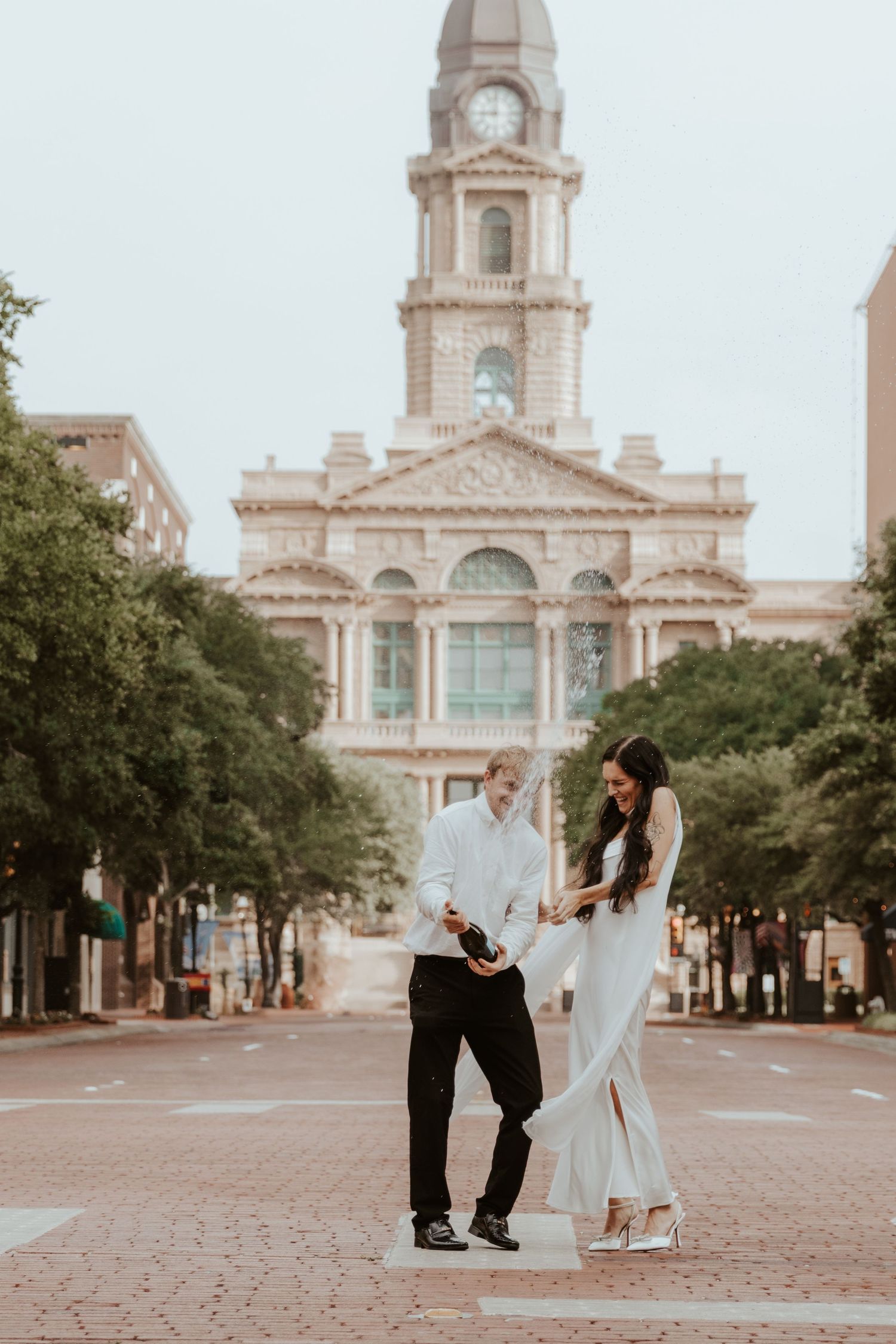 A couple poses for a romantic photo in front of a historic courthouse with a clock tower in Fort Worth, Texas at dusk.