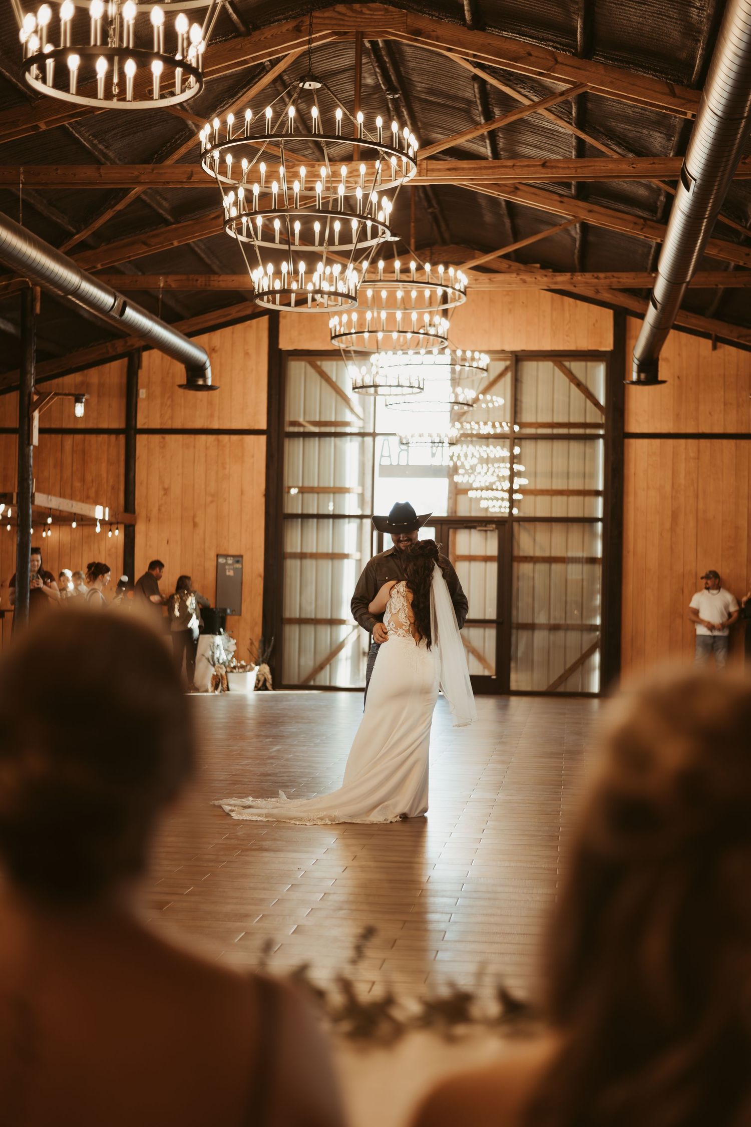 A wedding couple dances under elegant chandeliers in a rustic barn venue with wooden beams and warm lighting.