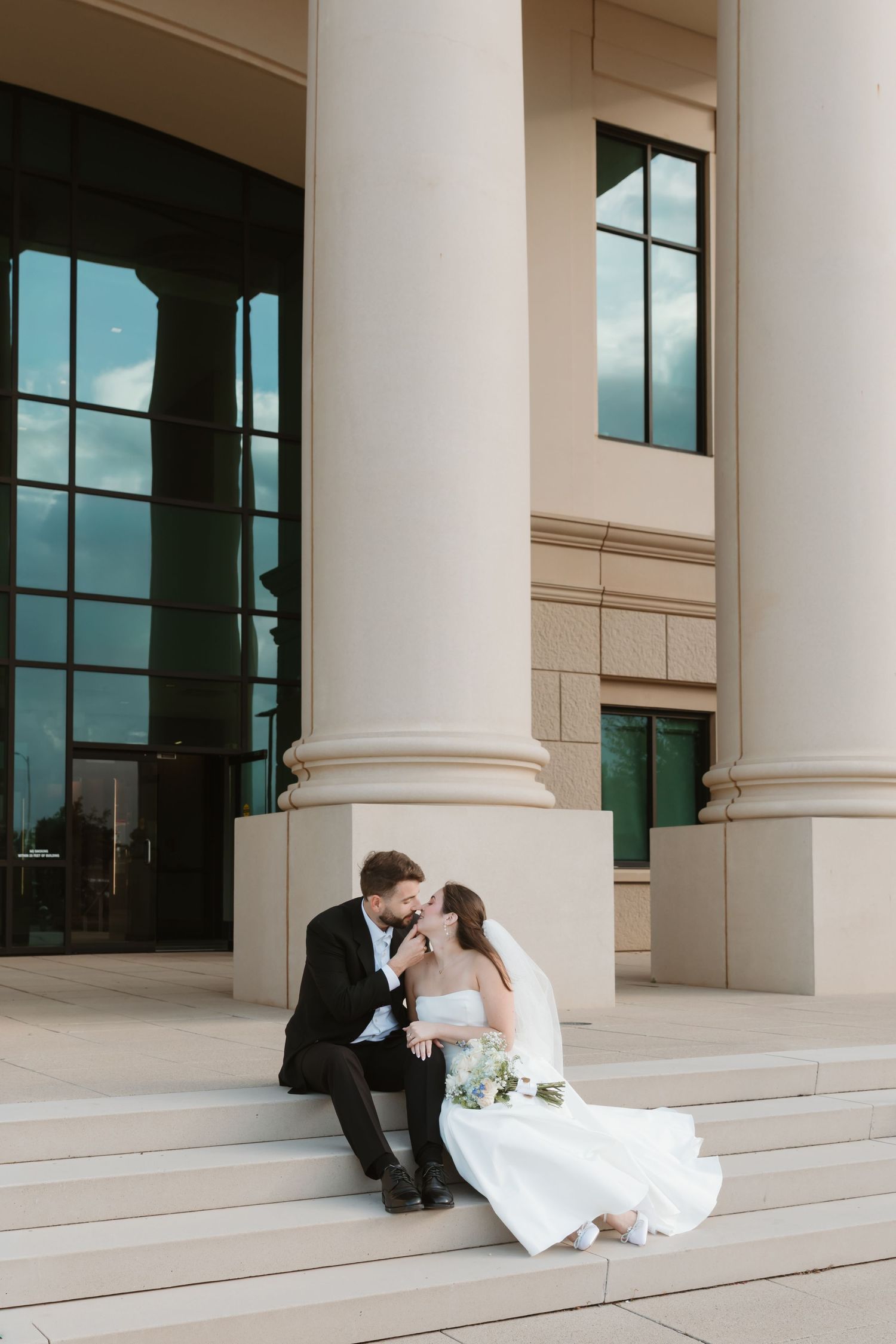 A romantic wedding couple shares a tender moment while sitting on elegant white stone steps of a columned building.