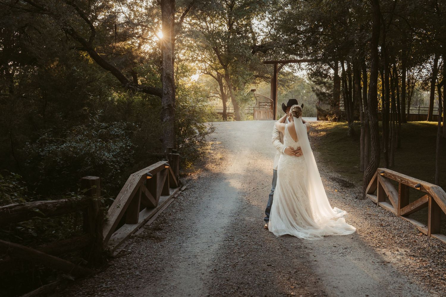 A bride in a white wedding dress stands on a rustic wooden bridge surrounded by sunlit trees during golden hour. Wedding Photographer DFW