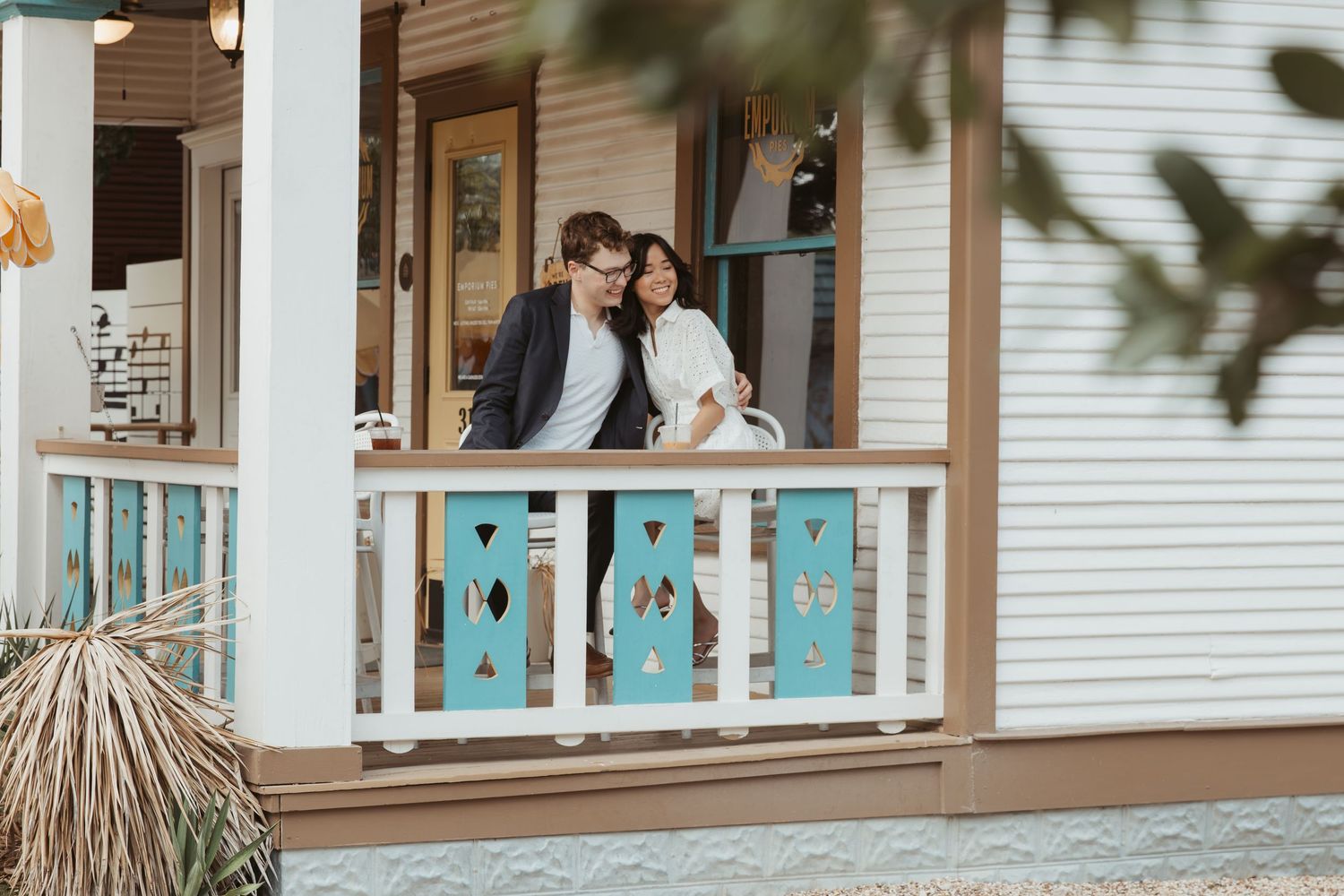 Couple embracing on a rustic white porch with turquoise doors and thatched palm decor in the background.