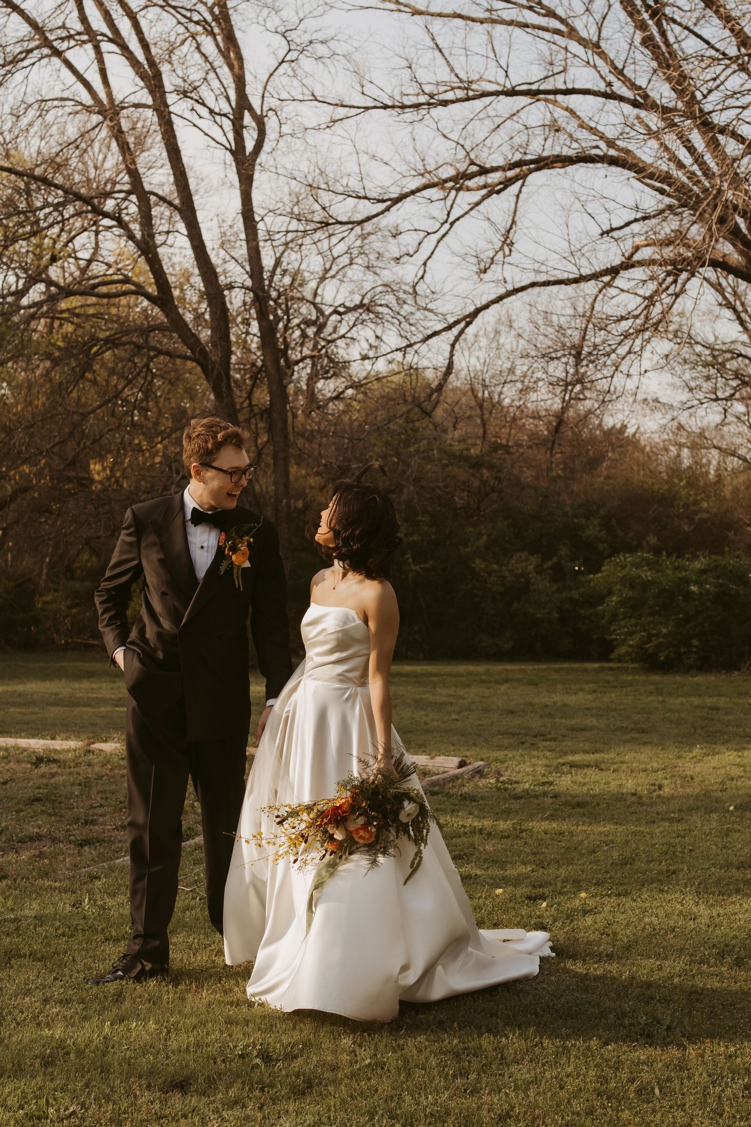Bride in white strapless gown holding bouquet stands with groom in black tuxedo outdoors among bare trees.