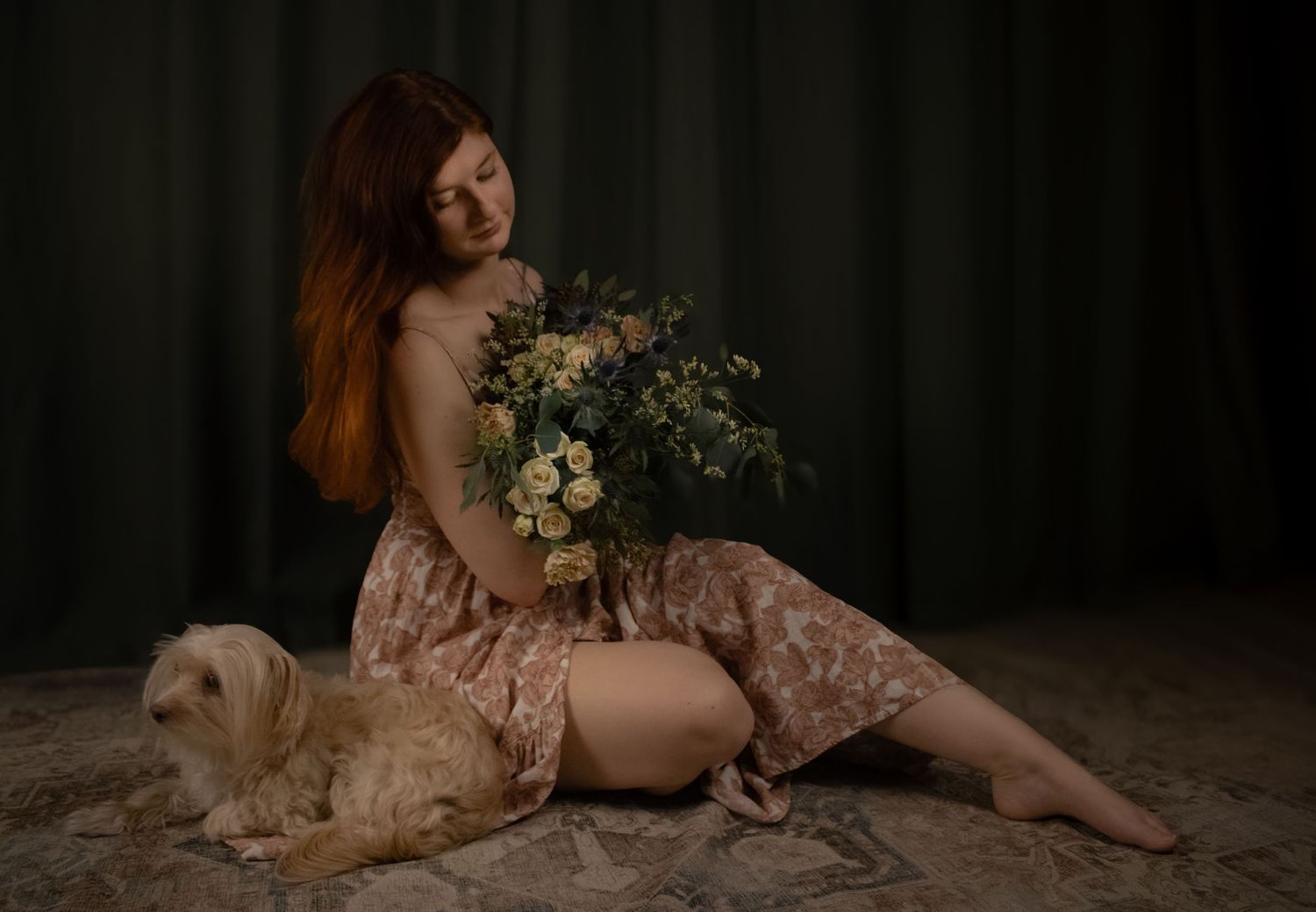 A woman in lace gown reclines on the floor with a bouquet of flowers and white dog in moody studio lighting.