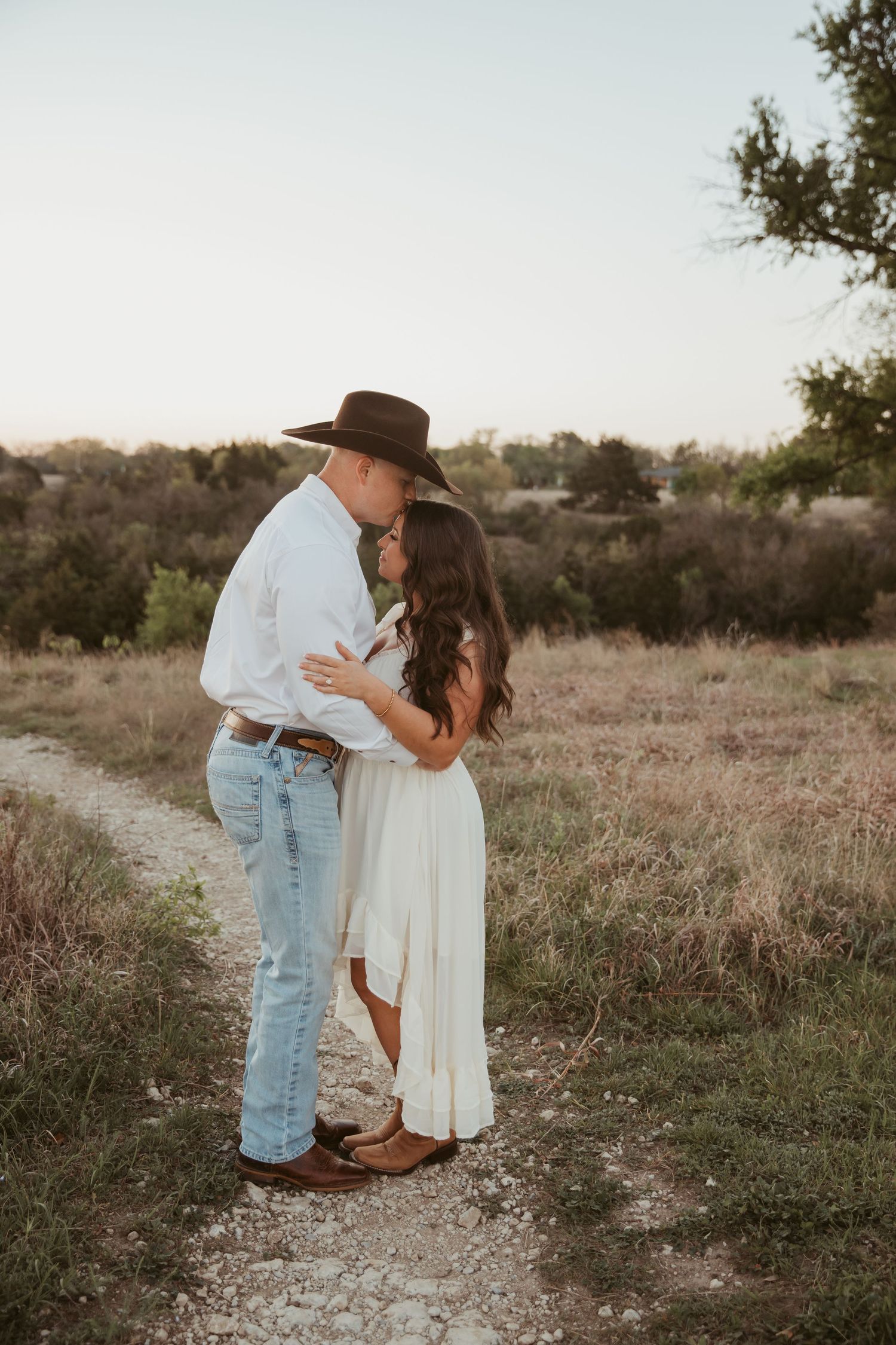 Couple sharing a romantic kiss on a country path, man in cowboy hat and jeans, woman in white dress, golden hour.