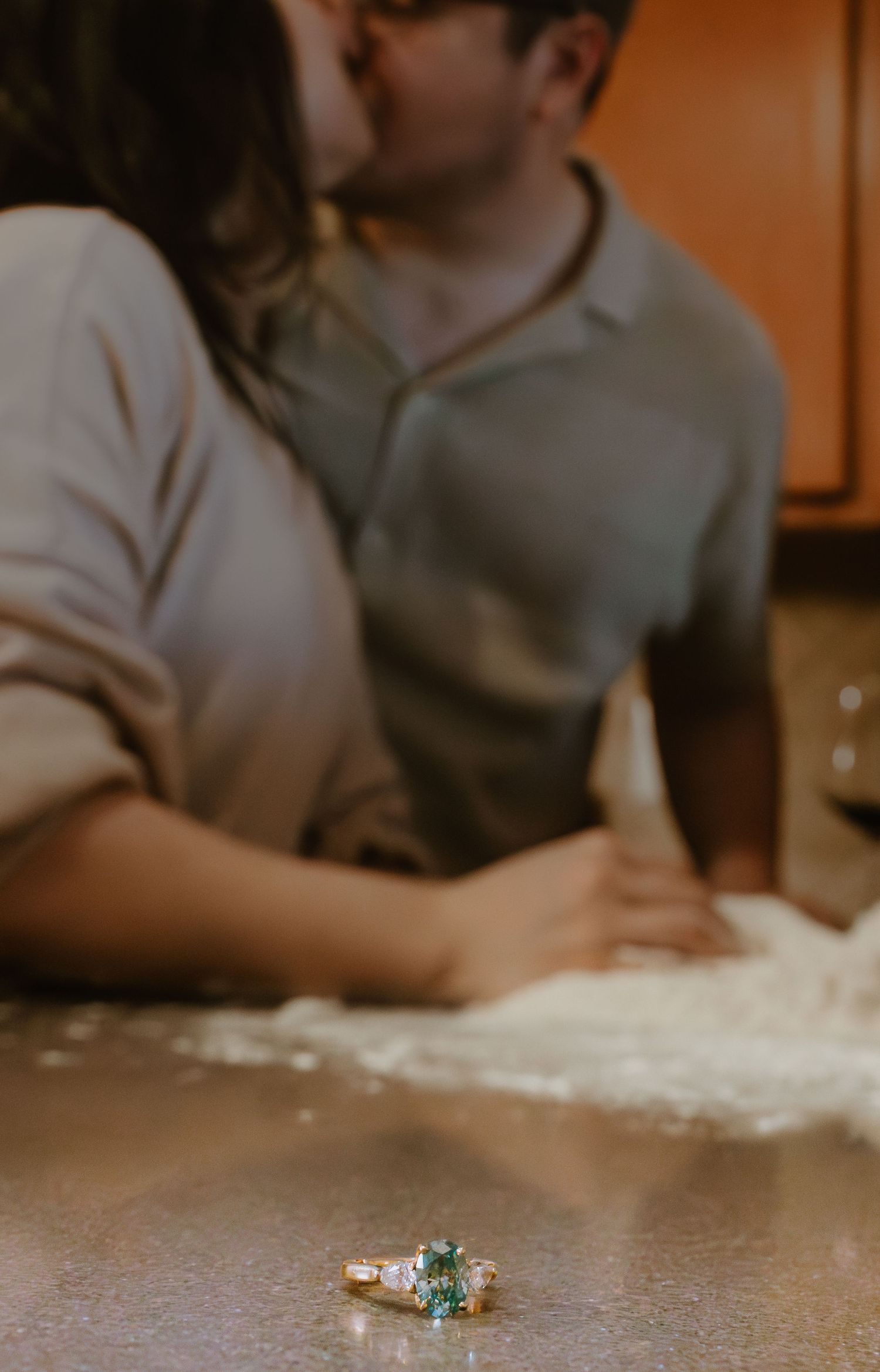 A teal engagement ring sits on a flour-covered surface while two people bake in the background.