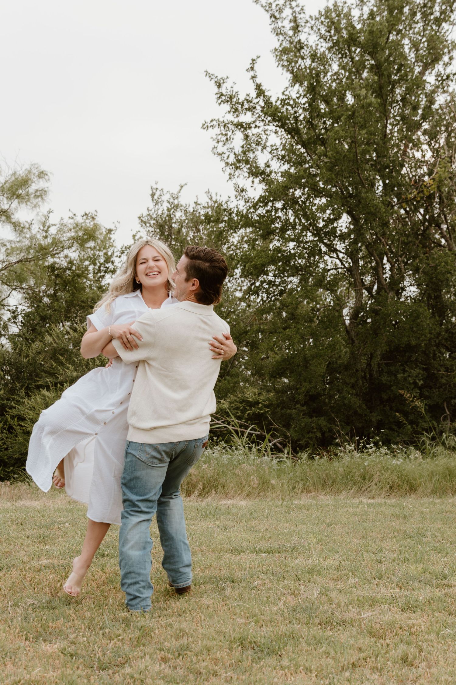 A couple shares a joyful embrace while dancing together in a grassy field with trees in the background.