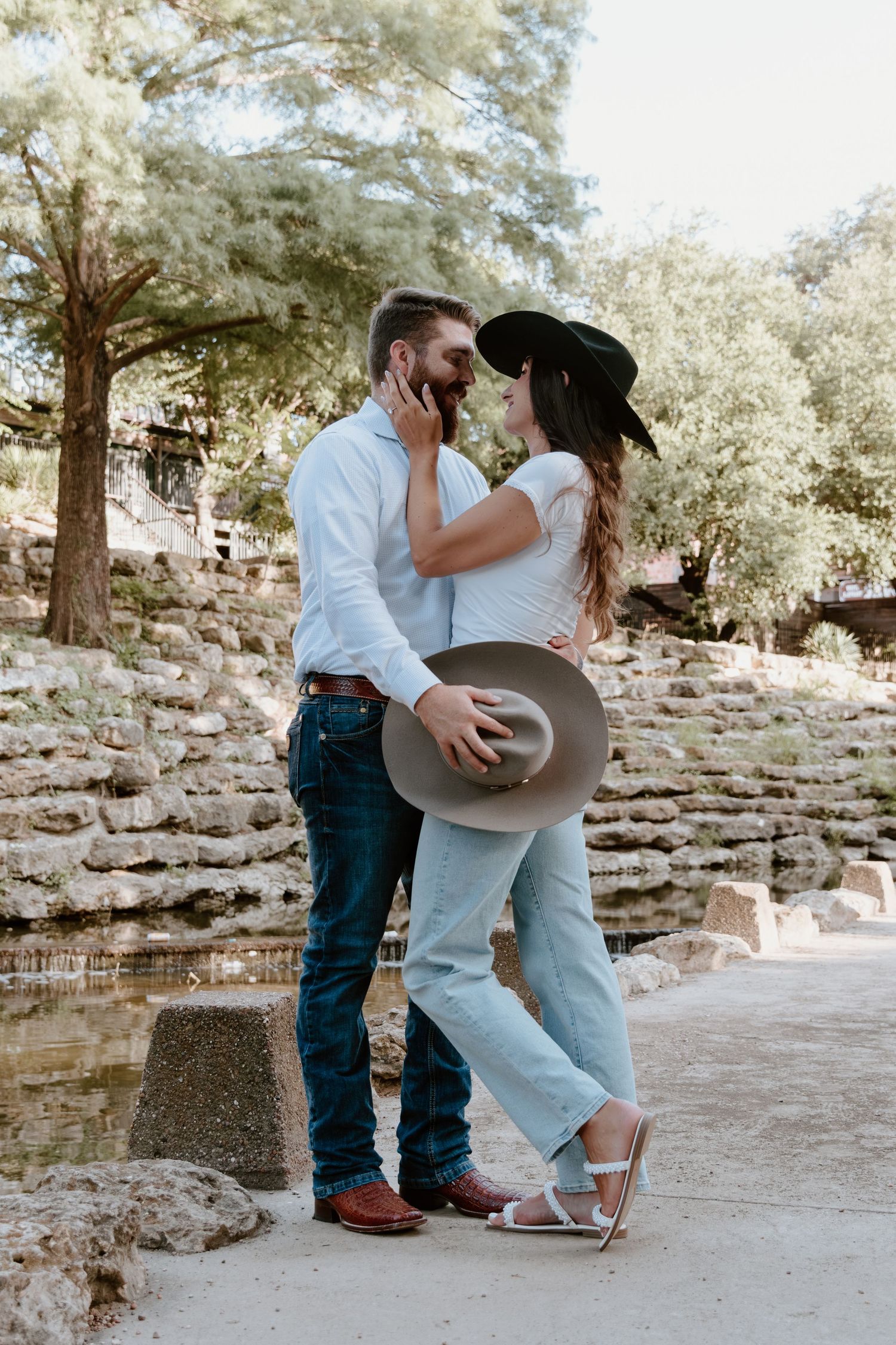 A romantic couple in western attire shares tender moments in a rustic outdoor setting with stone walls and trees.