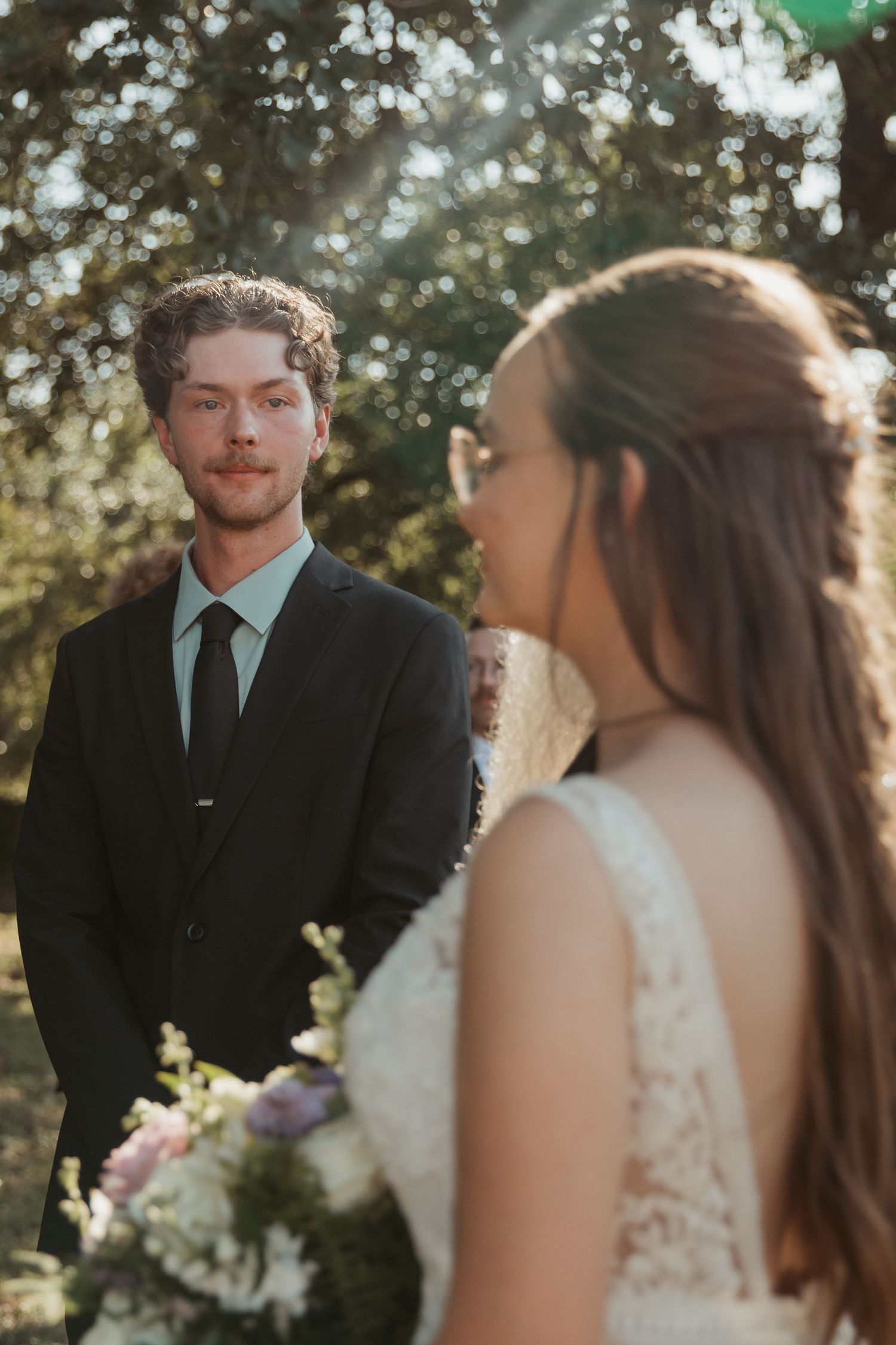Groom in black suit and green tie gazes at bride in lace dress during outdoor wedding ceremony.