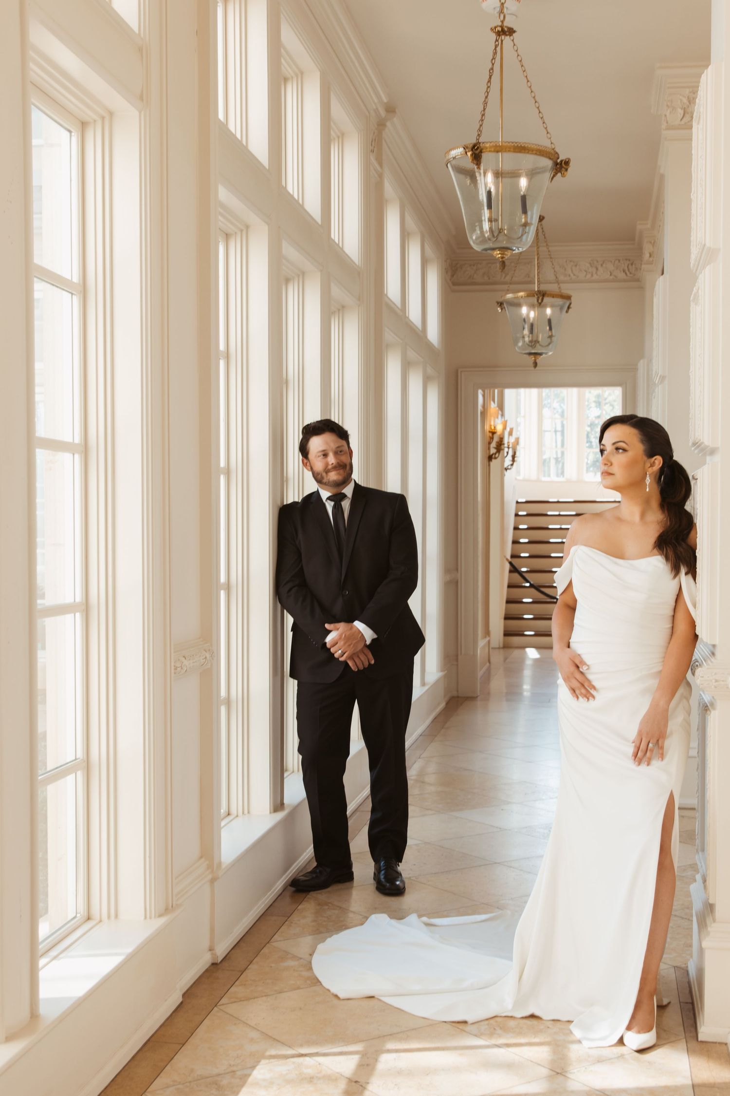 A bright sunlit corridor with white walls features elegant pendant lights and tall windows creating a dramatic atmosphere.