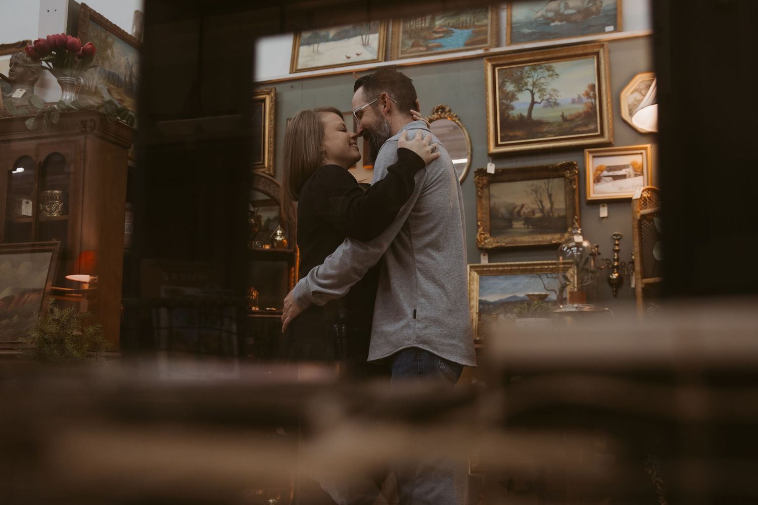 A romantic couple embraces in a dimly lit vintage room with framed artwork on the walls.
