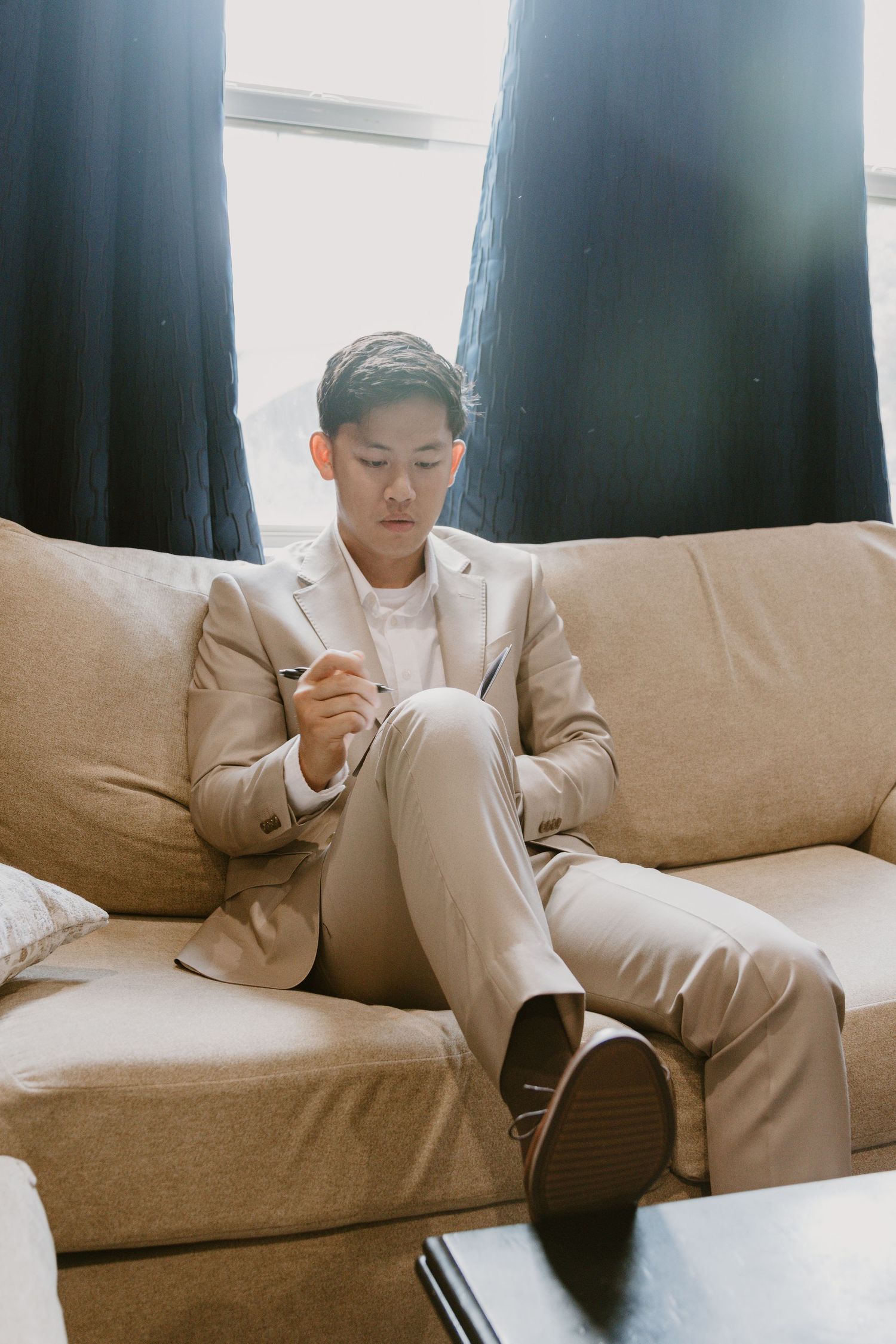 A person in a beige suit sits thoughtfully on a tan couch in an indoor setting.