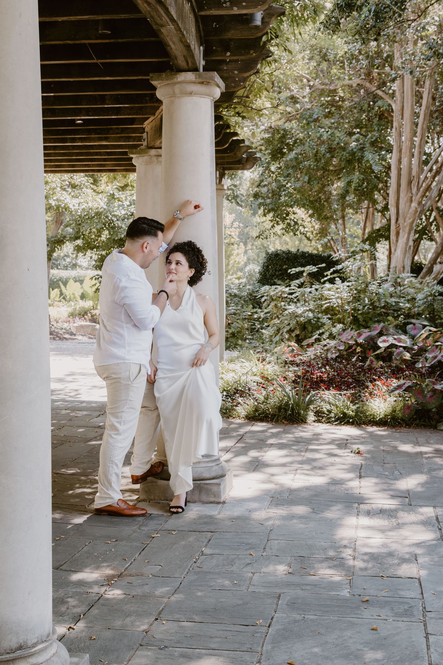 A couple in white outfits share a romantic moment under a columned porch with lush greenery in the background.
