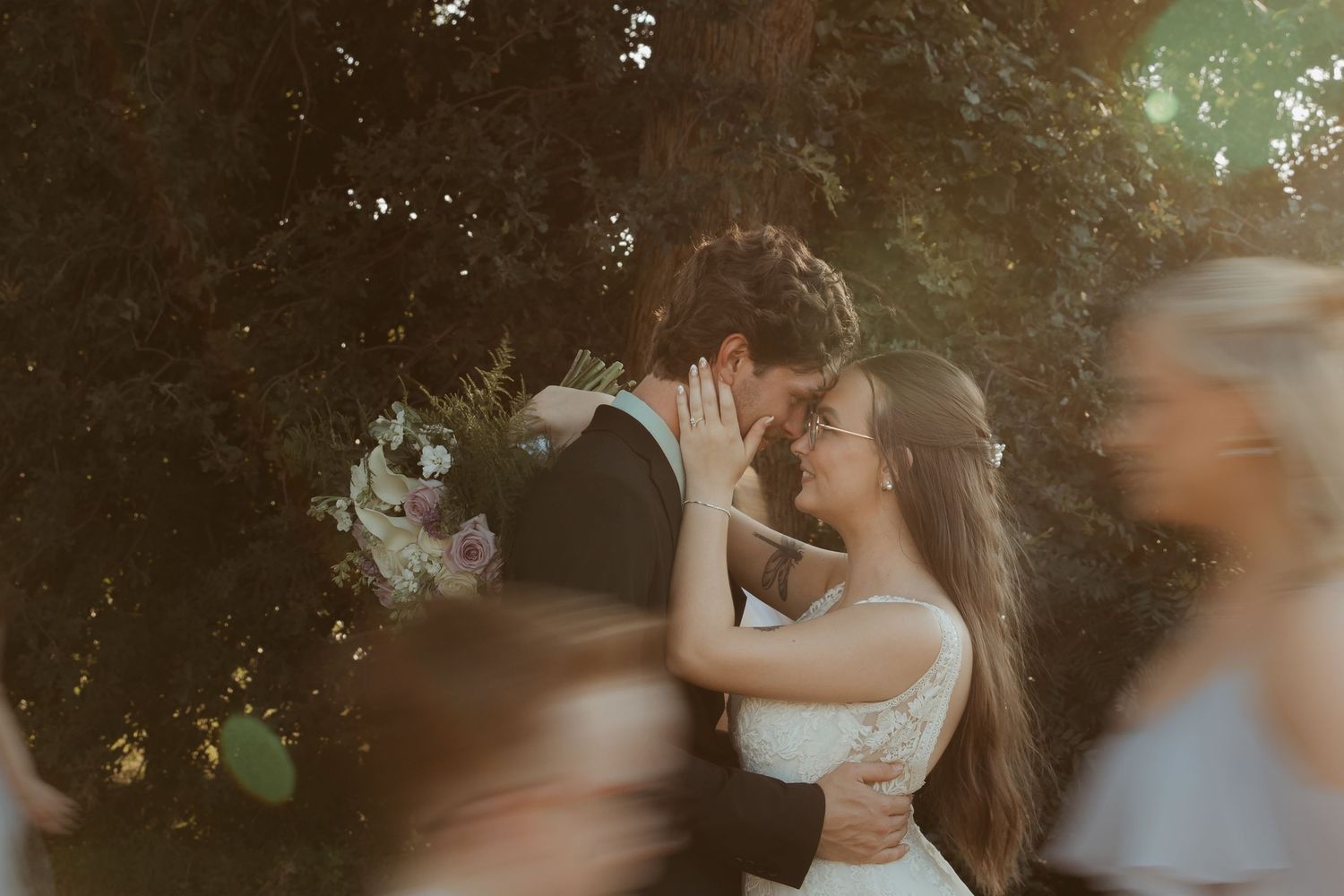 Bride and groom sharing a romantic kiss outdoors, surrounded by lush greenery and soft bokeh blur of wedding guests.