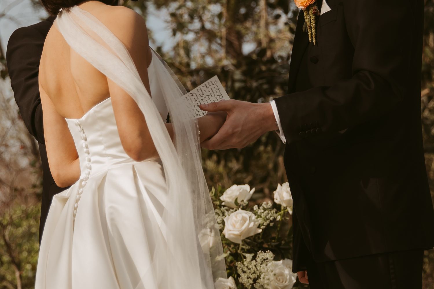 Bride in white strapless gown with veil stands beside groom in black suit during outdoor wedding ceremony.