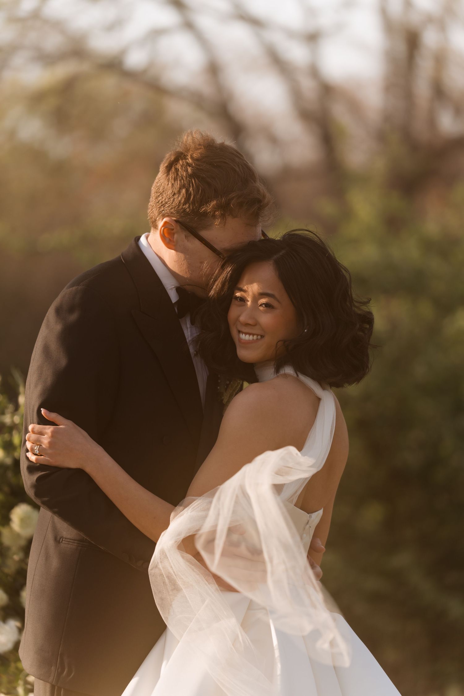 Bride in white tulle gown smiles as groom kisses her cheek during golden hour outdoor wedding portraits.