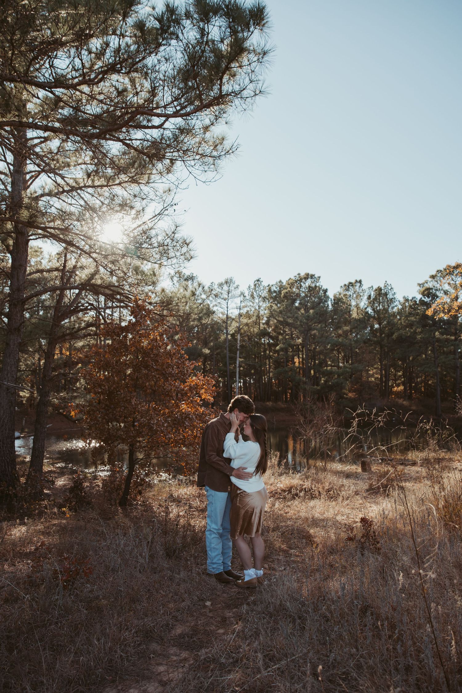 Two figures stand in a golden autumn field surrounded by pine trees at sunset, creating a romantic silhouette. engagement photos DFW