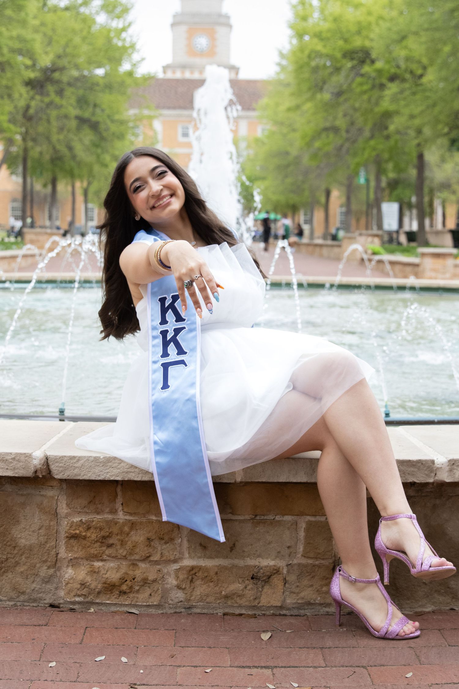 Graduate in white dress with light blue sorority sash poses by campus fountain on sunny spring day.