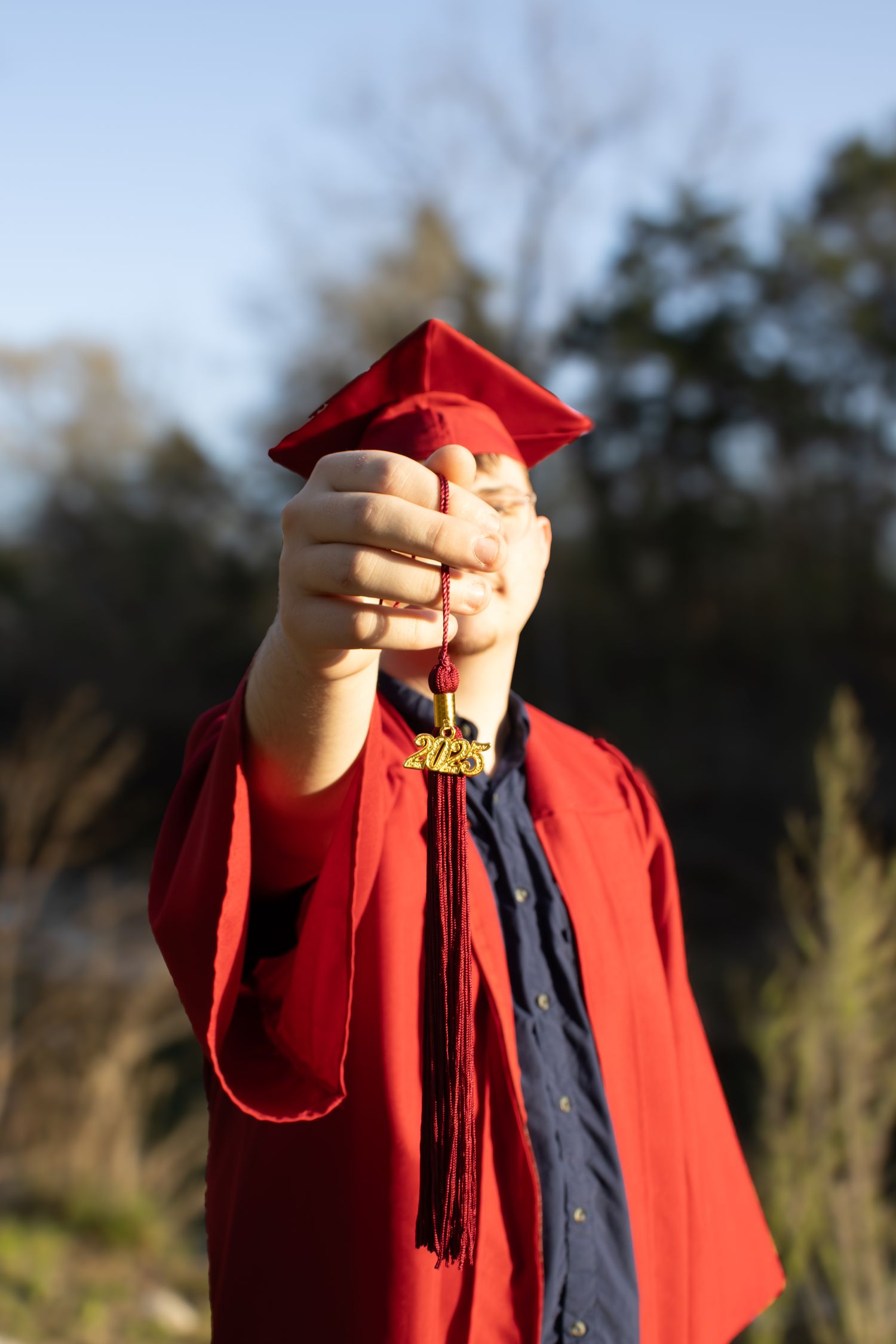 Graduate in red cap and gown holds up graduation tassel against blue sky and trees in outdoor setting.