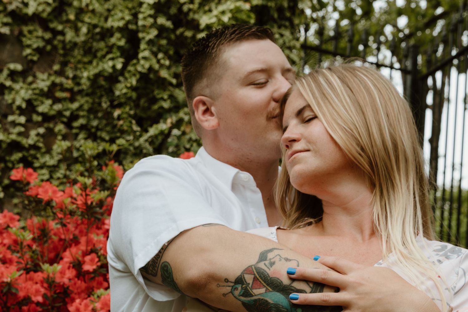 A couple share an intimate moment among flowering bushes as they pose for an engagement photo outdoors.
