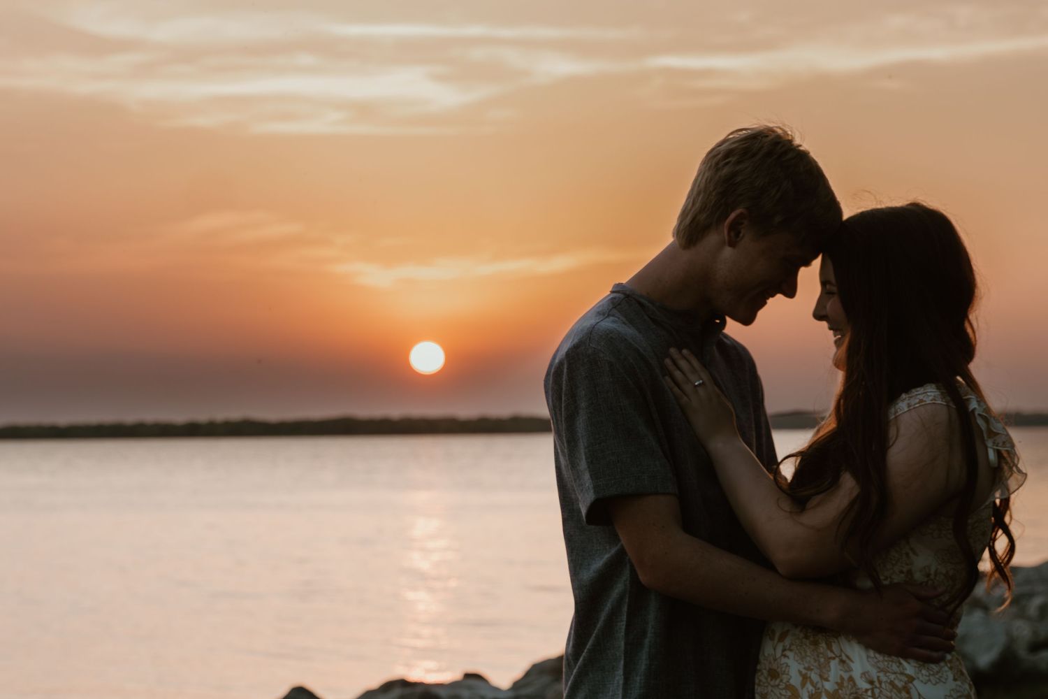 A silhouetted couple embraces during a romantic sunset by the water with an orange sky and glowing sun.