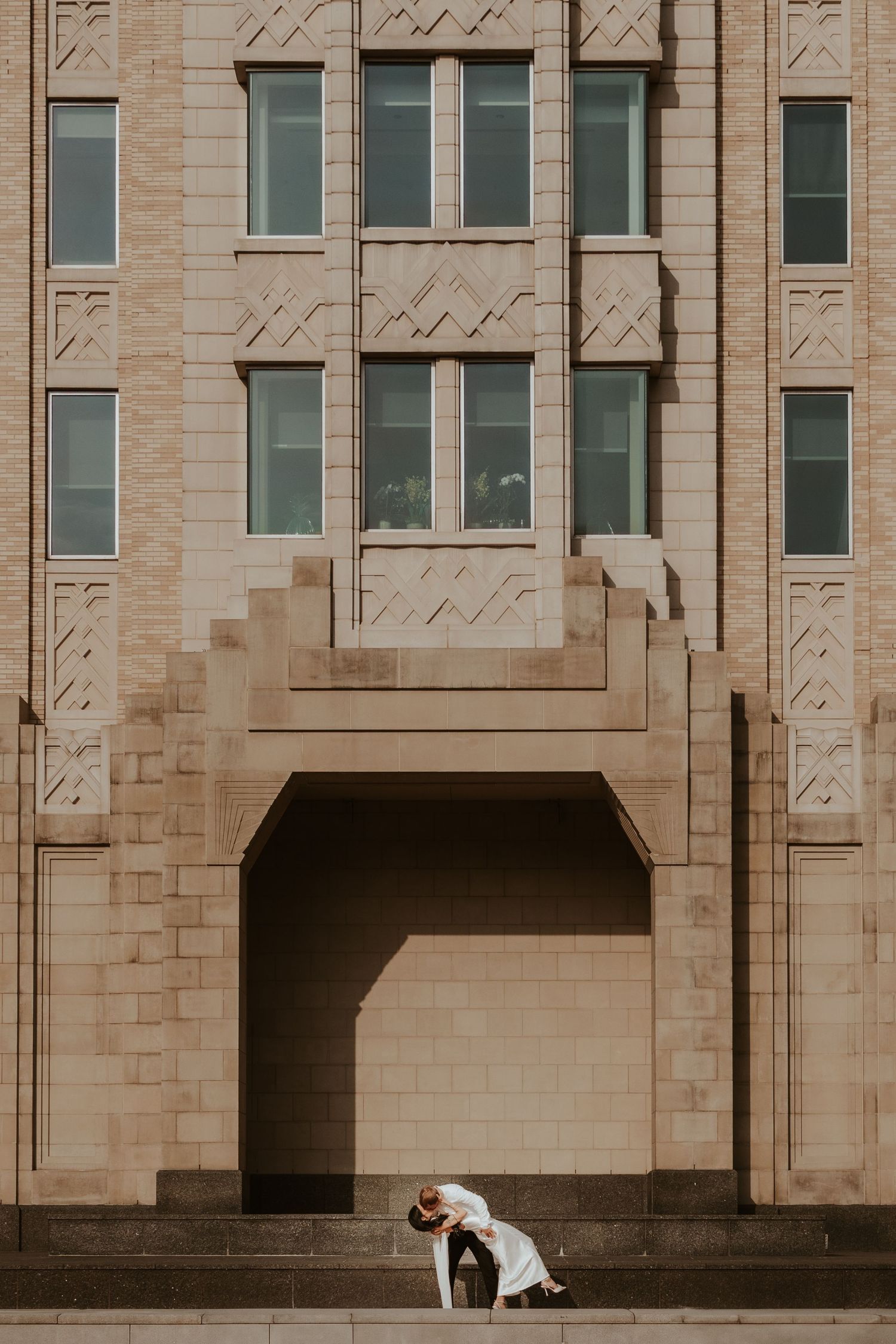 Two people in elegant attire share a romantic dip pose against a grand Art Deco style building exterior.