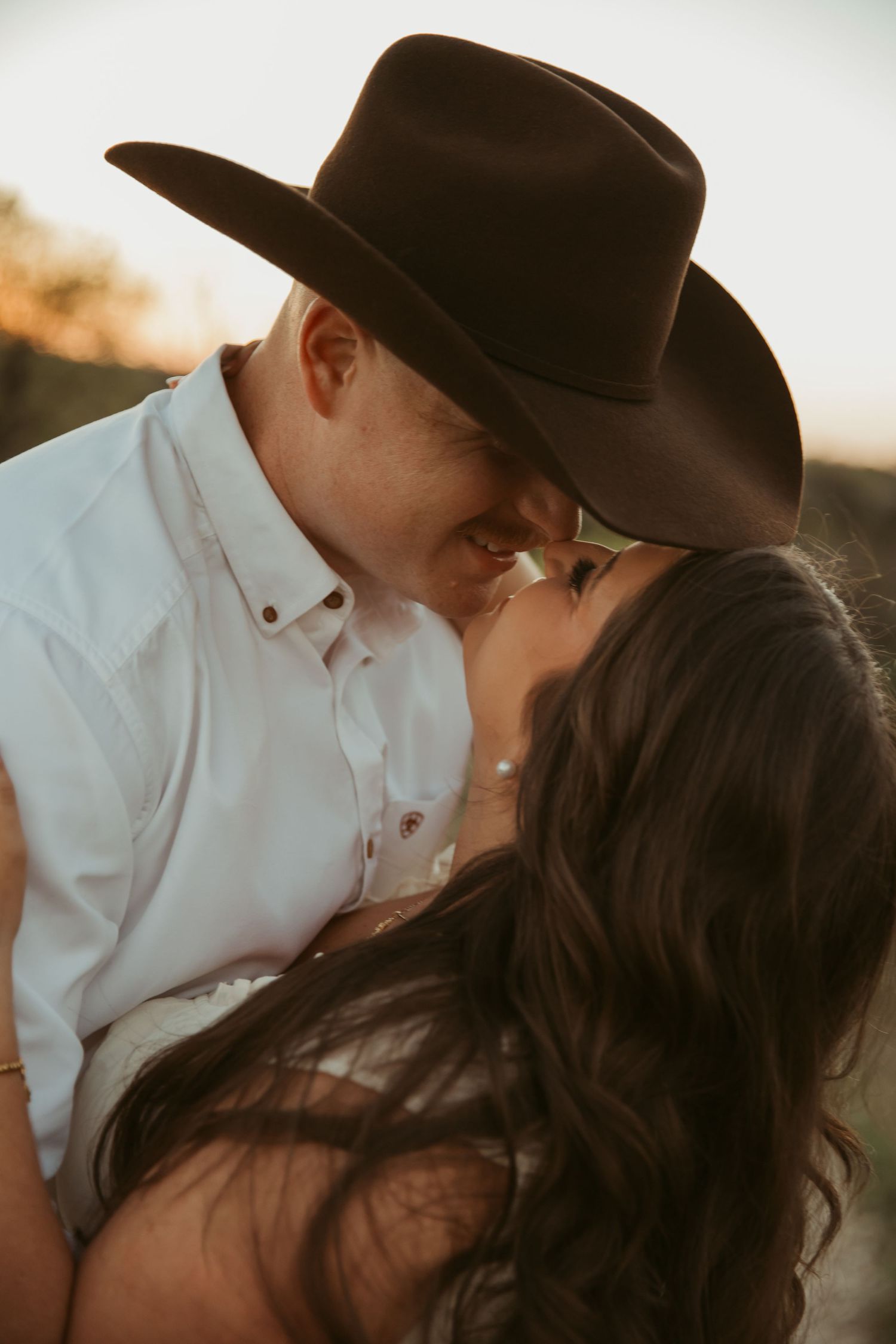 Couple sharing a romantic kiss outdoors at golden hour, man wearing a black cowboy hat and white shirt.