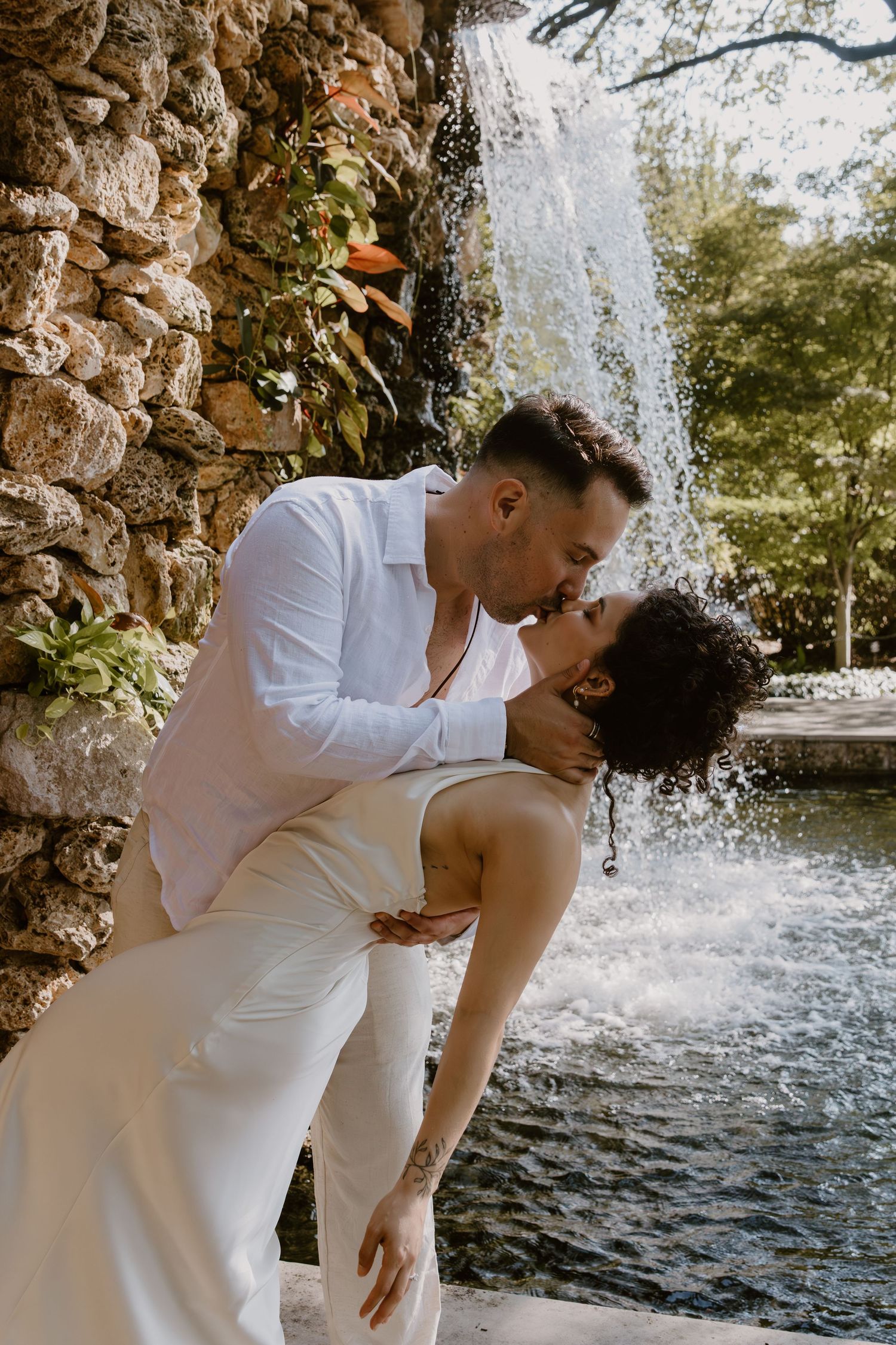 A romantic couple in white outfits share an intimate moment in front of a cascading waterfall.