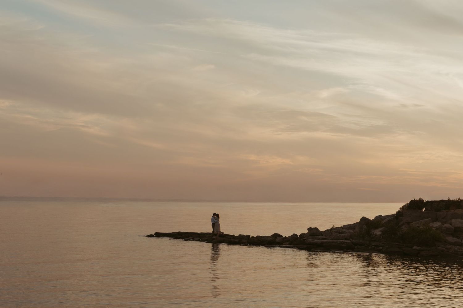 Lighthouse silhouette on rocky shore during golden sunset over tranquil waters.
