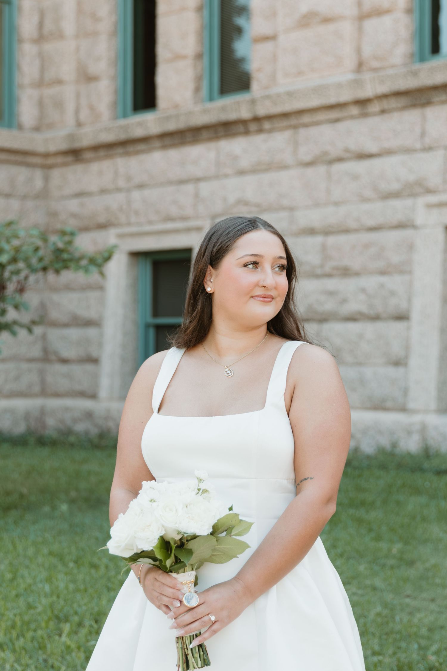 A person in a white wedding dress poses outdoors against a stone building wall holding a bouquet of white flowers. Wedding Photographer DFW