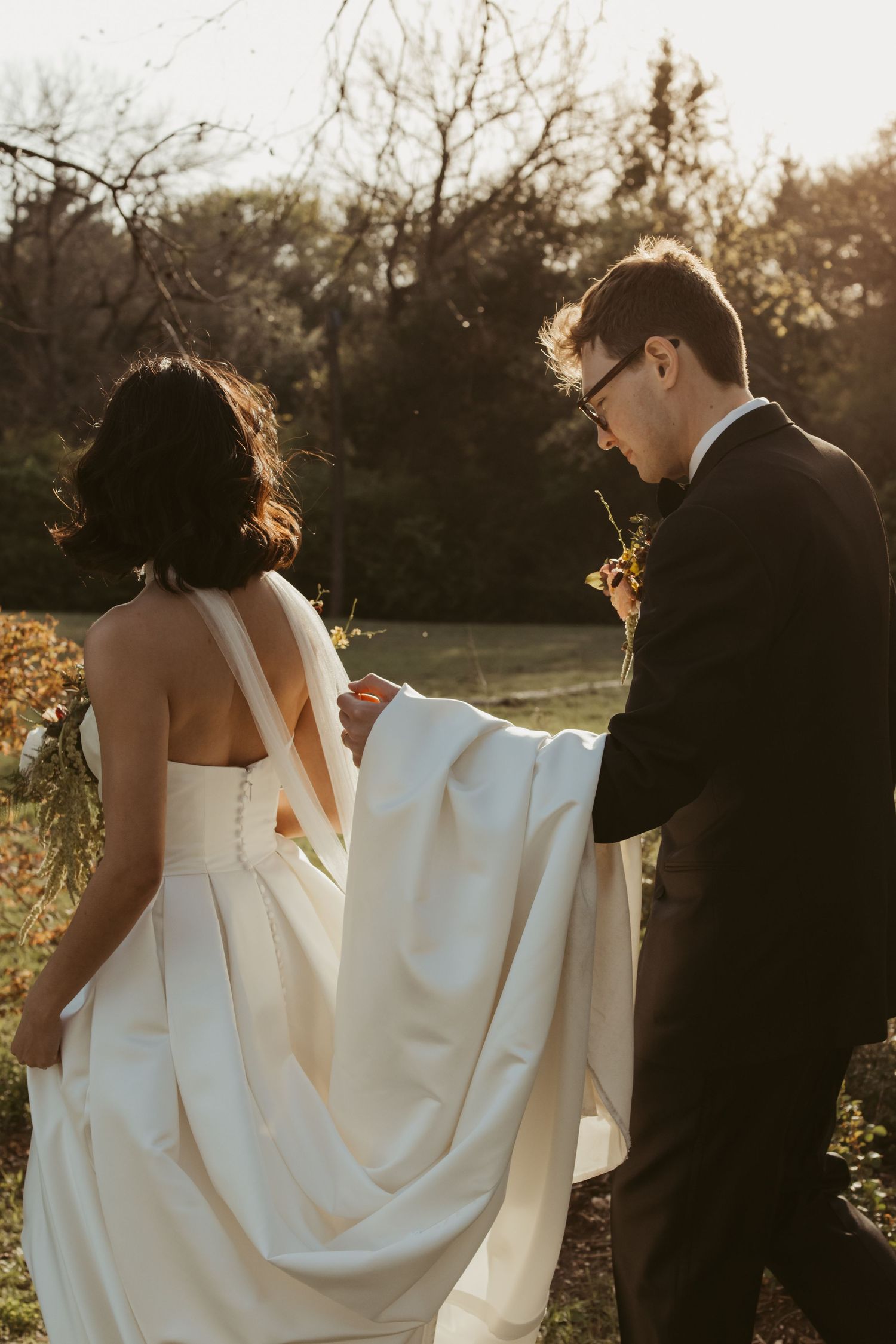 Groom in black tuxedo carries brides white satin gown train during golden hour outdoor wedding.