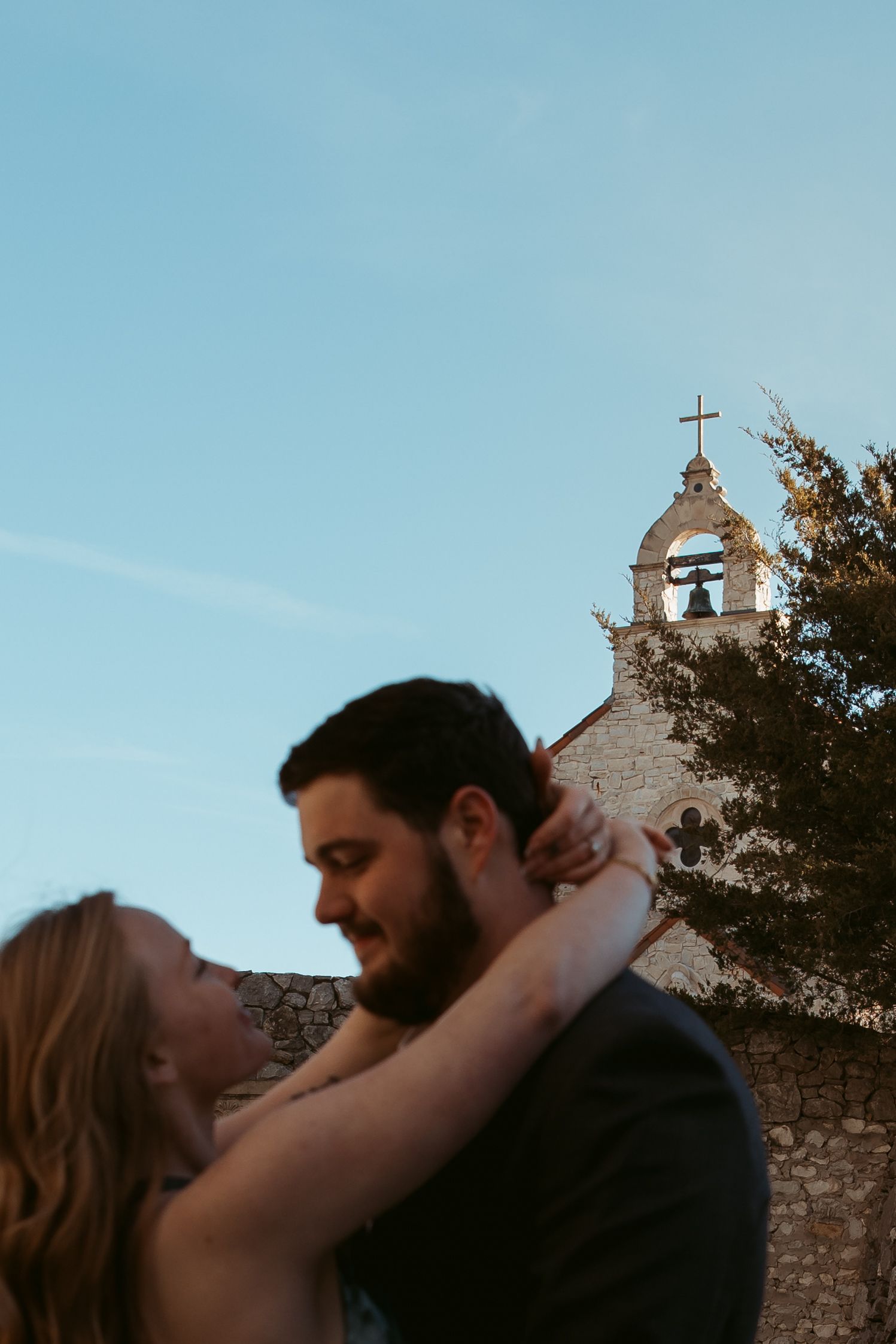 Silhouetted couple embraces against blue sky with historic church spires in background.