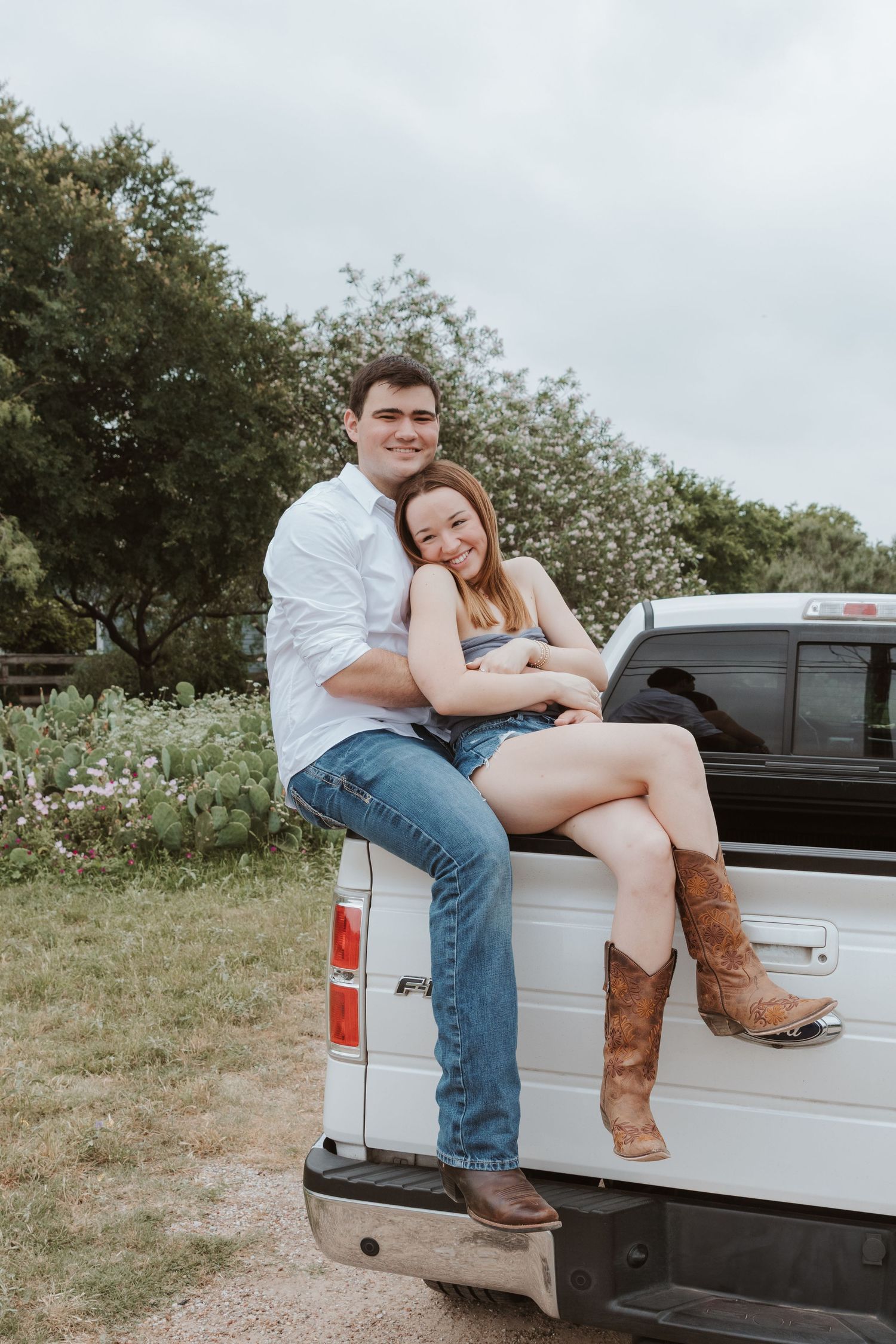 A couple sits together on the tailgate of a white pickup truck in a casual outdoor setting with trees in the background.