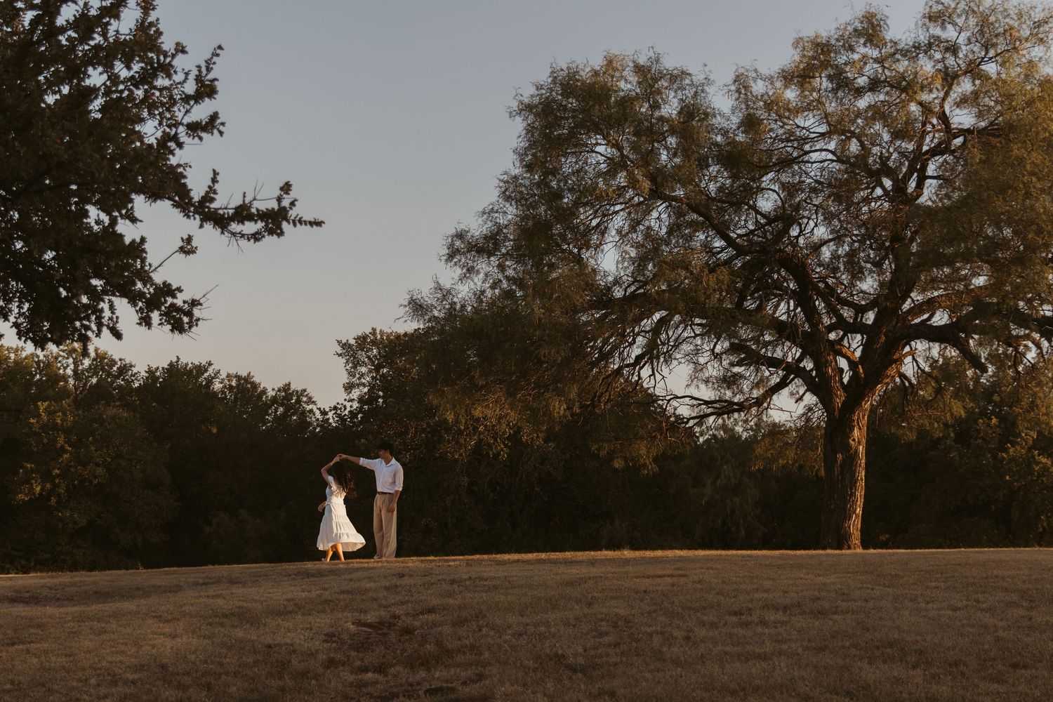 A couple in wedding attire stands on a hilltop during golden hour with trees and warm sunset lighting in the background.