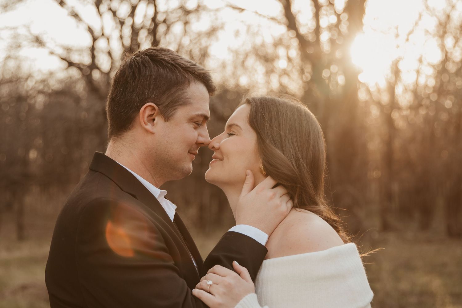 A couple shares a romantic golden hour moment, nearly kissing outdoors, the woman in an off-shoulder wrap and man in a suit.