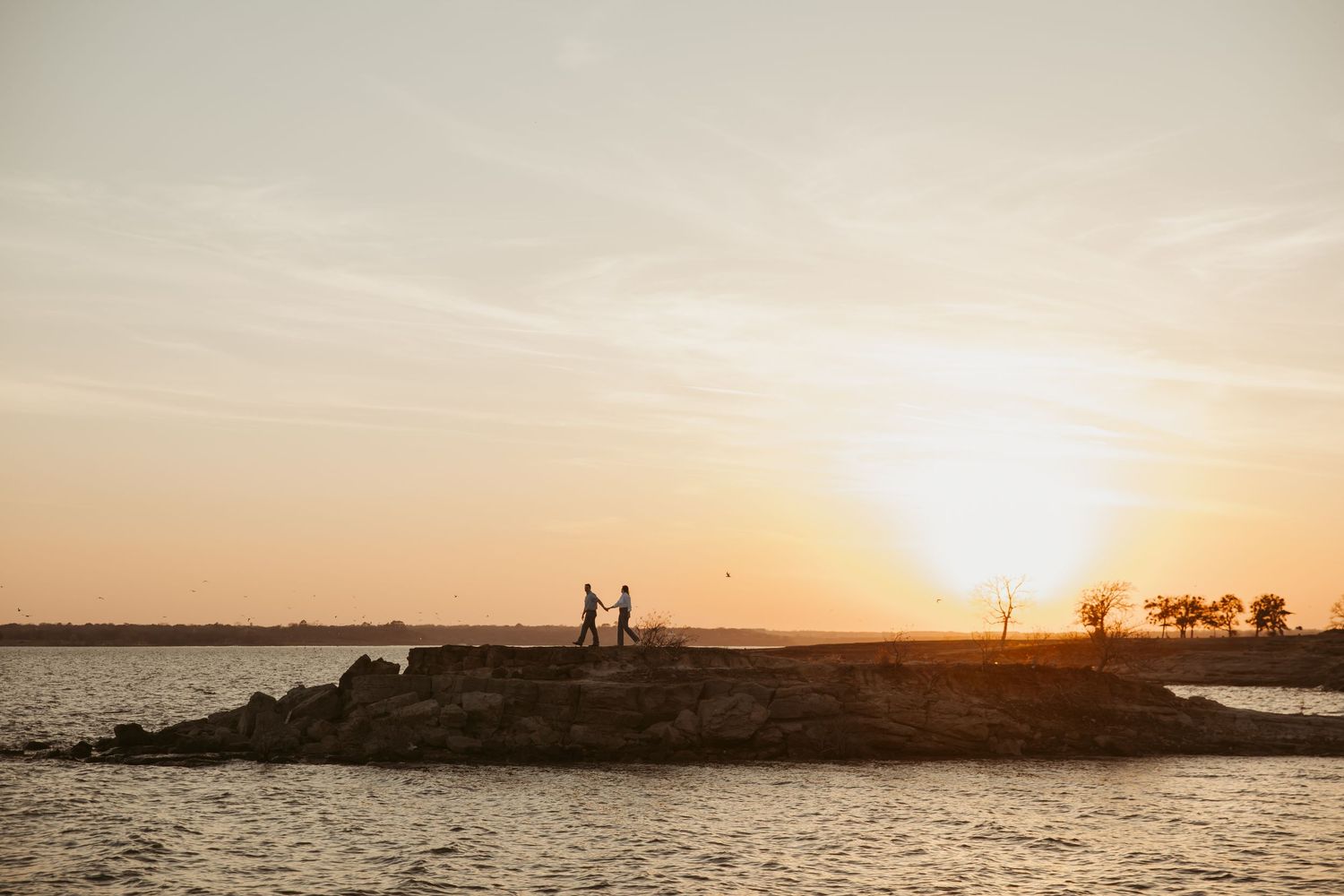 Silhouette of couple holding hands on rocky shoreline at golden sunset with calm water reflections.