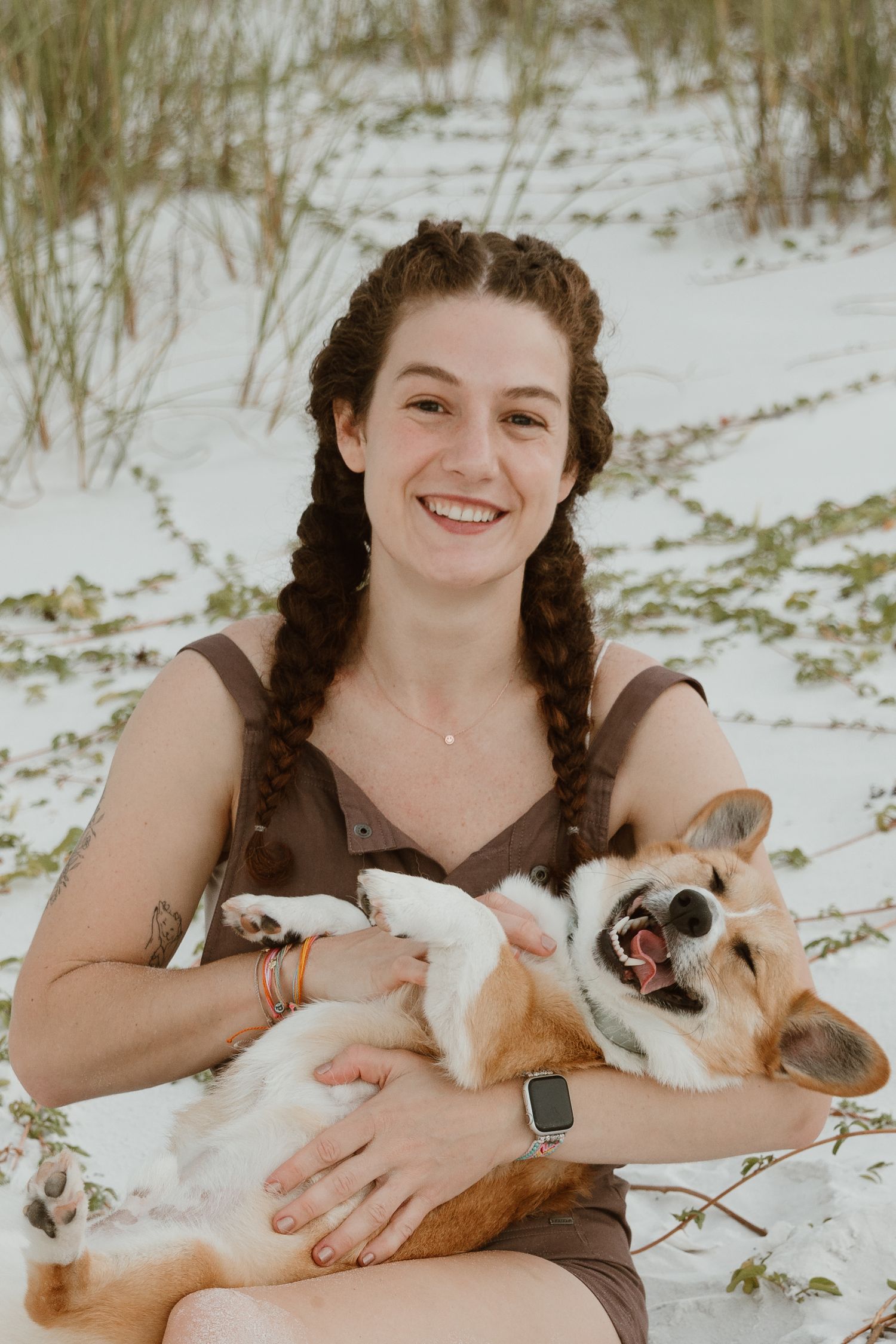 Smiling young woman with braided hair holding a happy Corgi dog on a white sand beach.