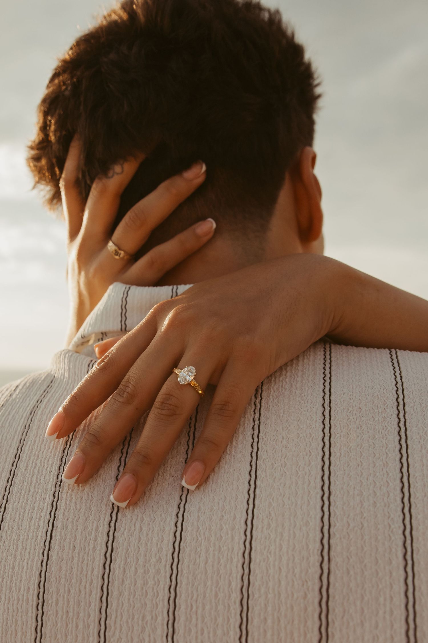 A woman's hand with a diamond engagement ring rests on a man's shoulder during a romantic outdoor photo shoot.