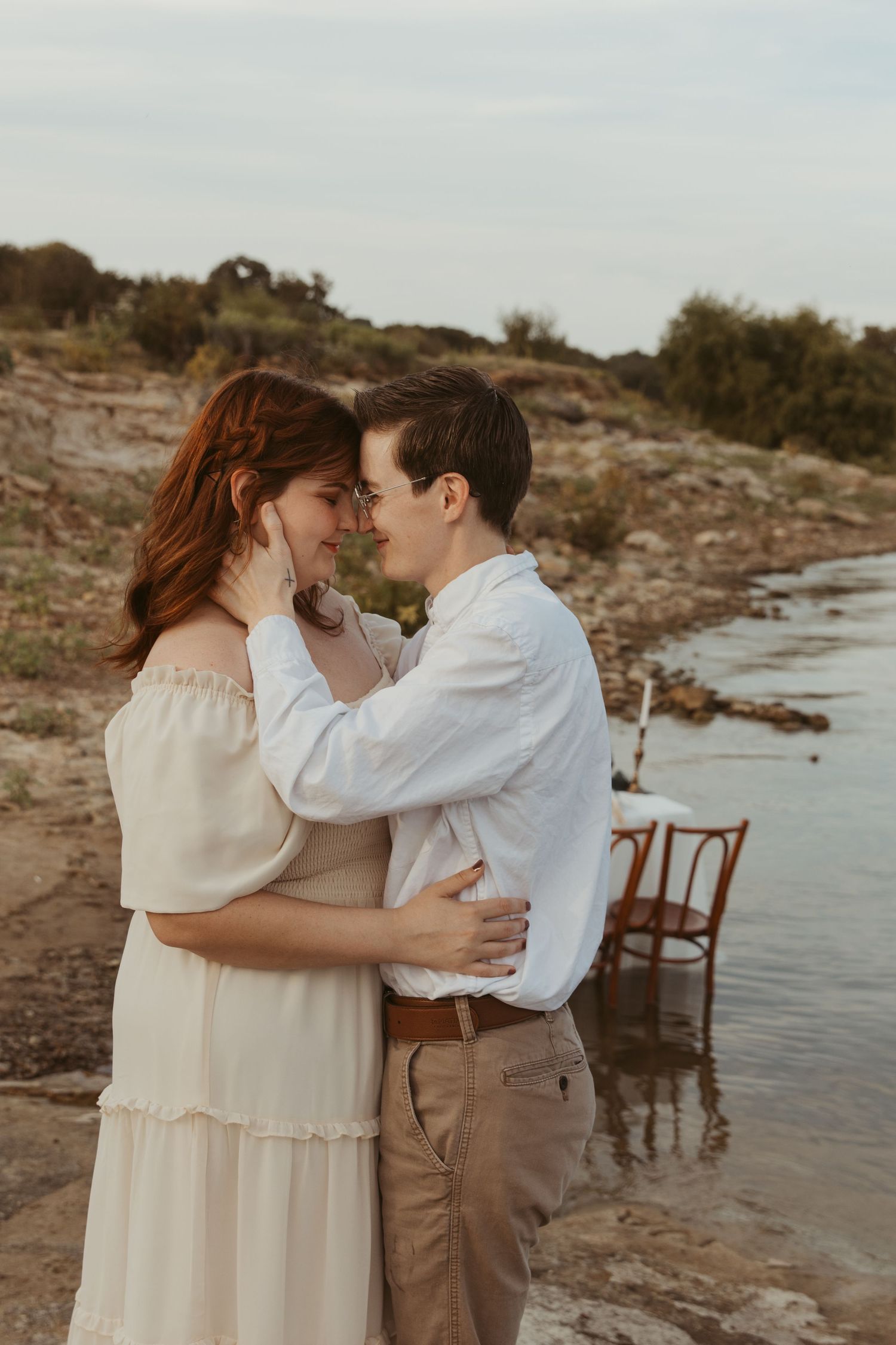 Couple in vintage-inspired attire share intimate moment by the water with wooden chairs nearby.
