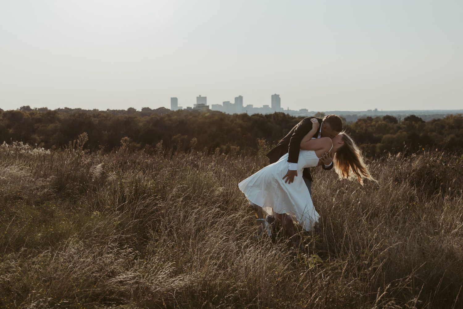 A person in a white dress twirls gracefully in a golden field at sunset with a city skyline visible in the distance. Engagement photographer