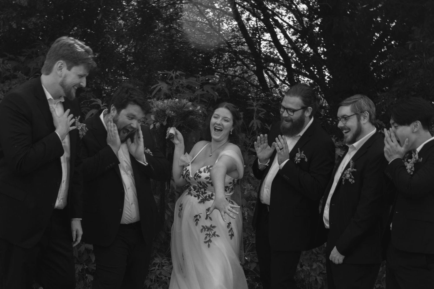 A black and white wedding party photo showing a candid moment of laughter among the bride and groomsmen outdoors. Wedding Photographer DFW
