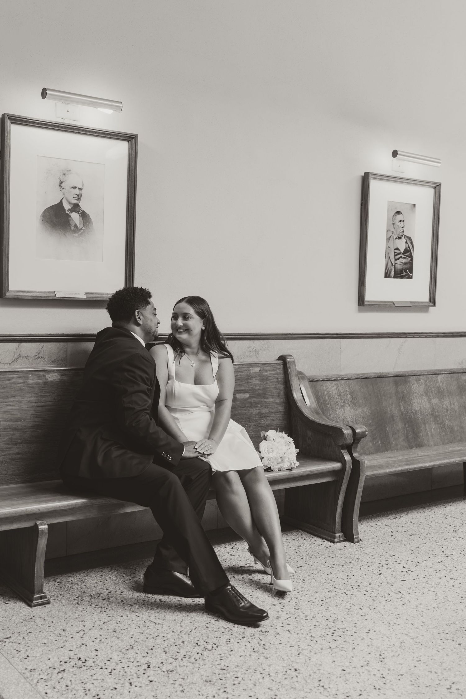 A couple sits together on a wooden bench in a hallway with black and white photography. Wedding Photographer DFW