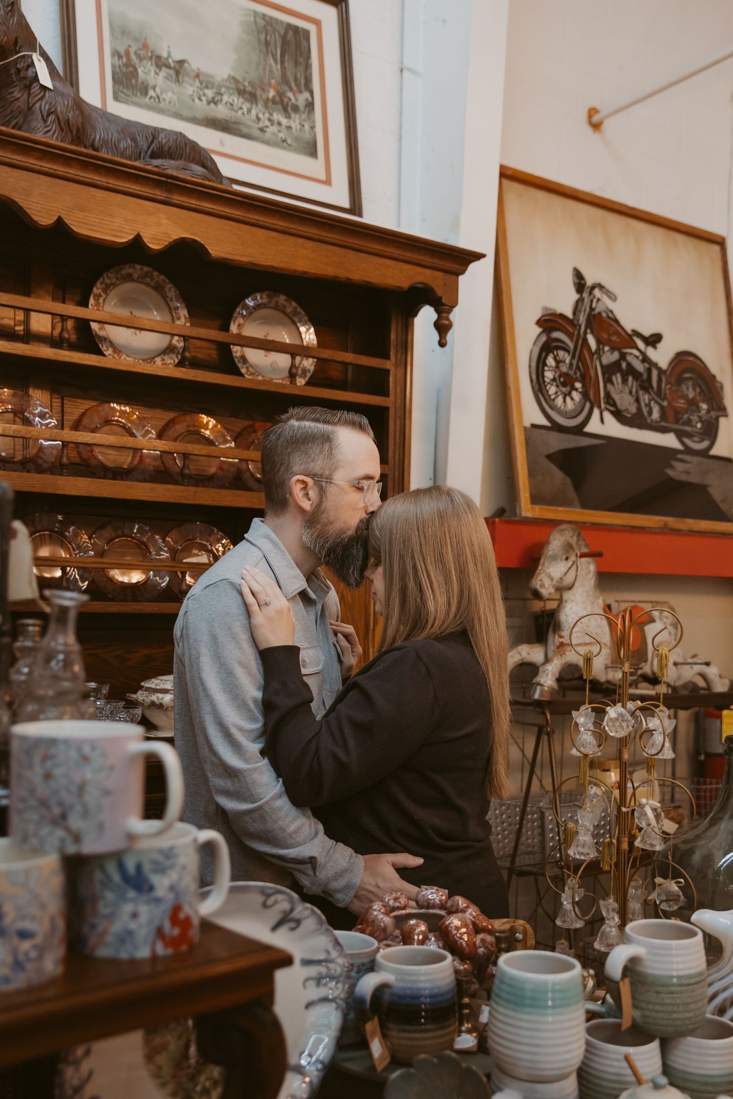 Couple sharing intimate moments in an antique shop filled with vintage plates and decorative items displayed on wooden shelves.