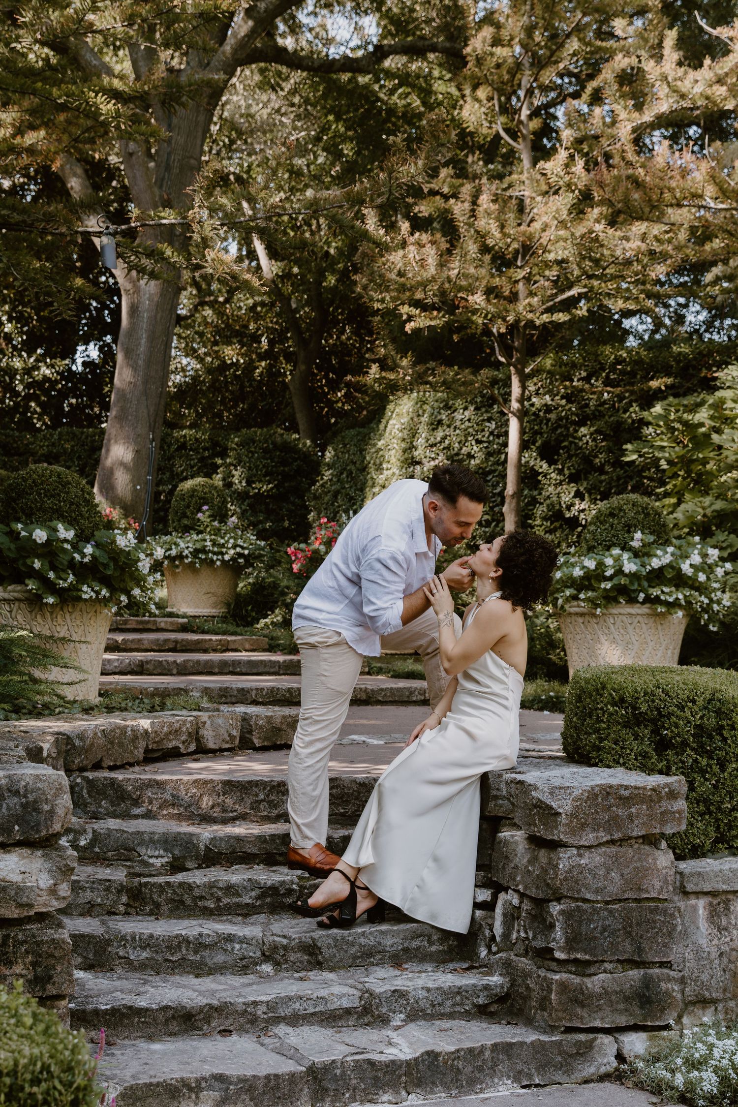 A couple in white attire shares intimate moments on stone steps in a garden setting.