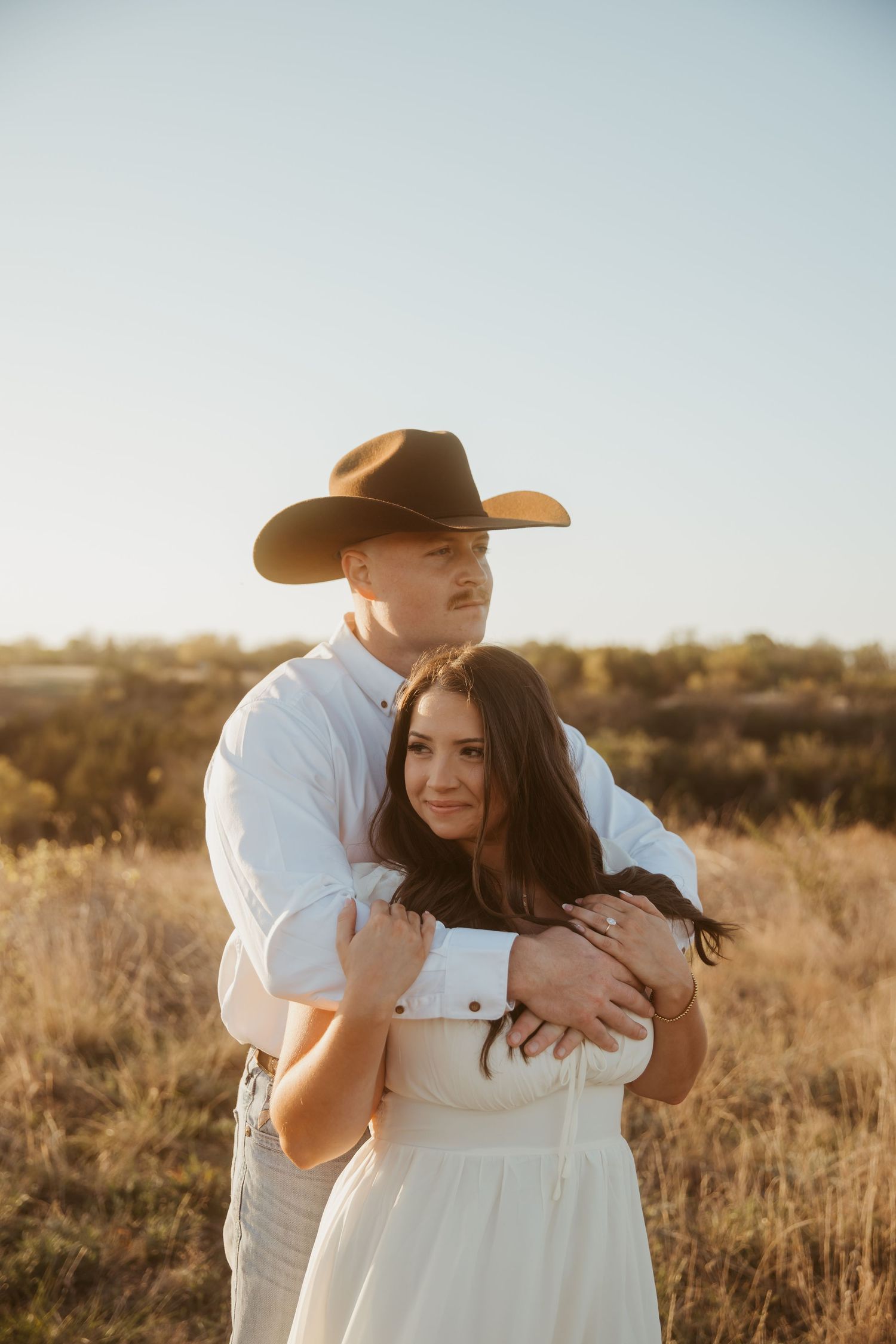 Couple embracing in golden field, man in white shirt and cowboy hat hugging woman in white dress at sunset.