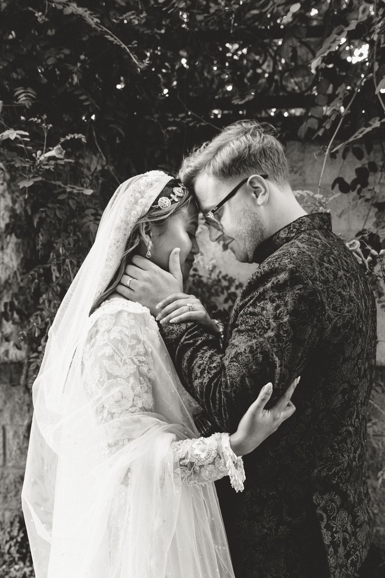 A romantic black and white wedding photo of a couple sharing an intimate moment outdoors among foliage.