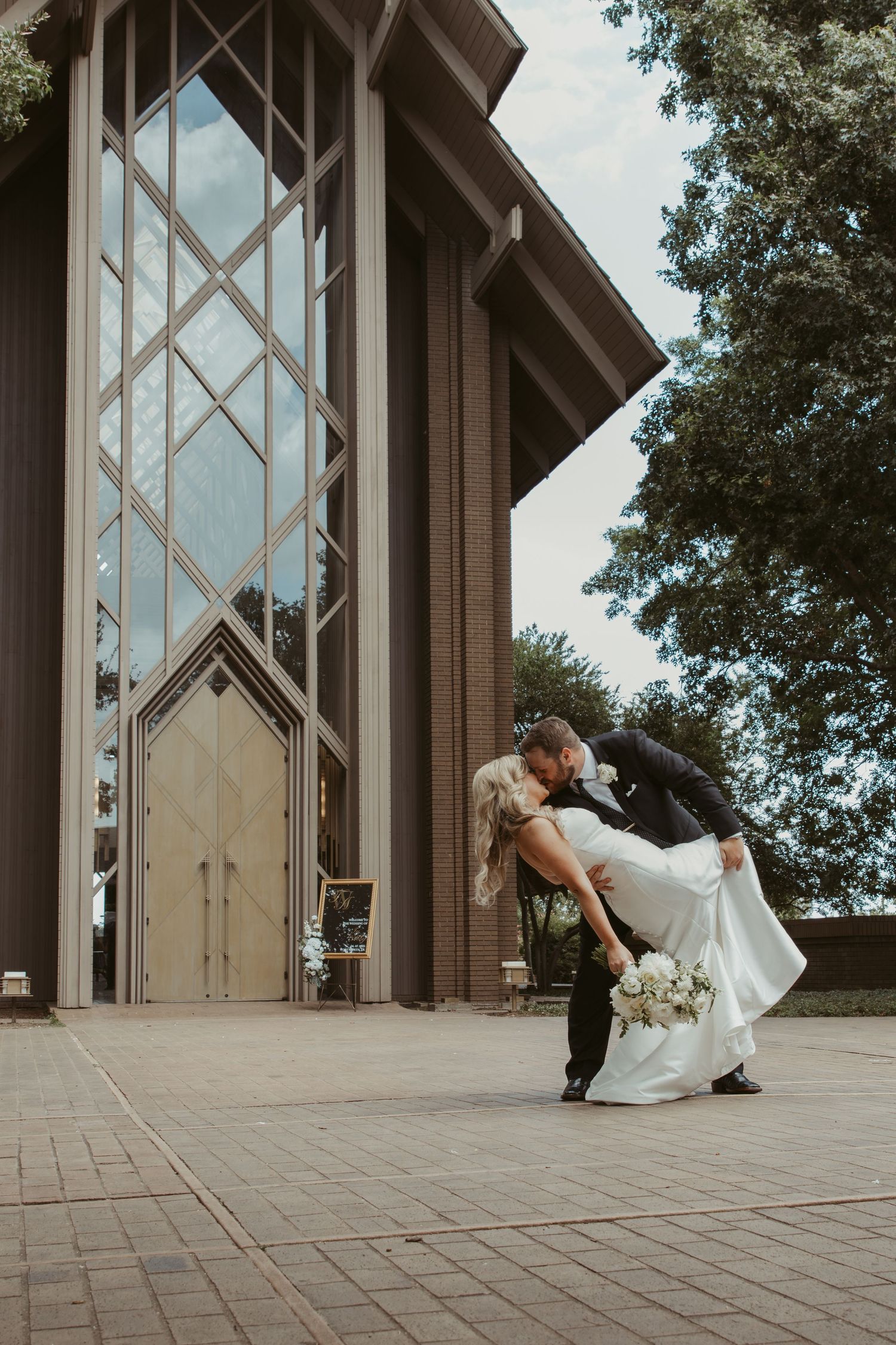 Low angle view of a couple in wedding attire lying on a concrete floor near architectural pillars and windows.