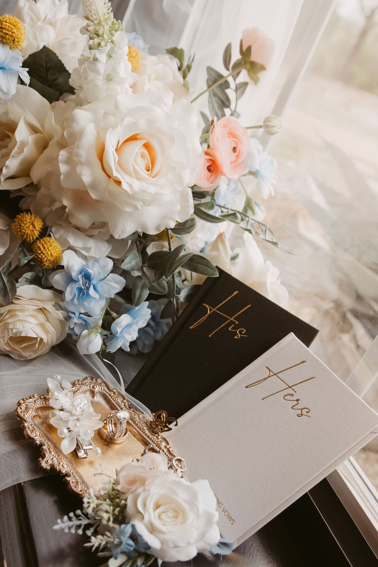 Wedding flat lay with His and Hers vow books, gold jewelry tray, rings, and a bouquet of white, pink, and blue flowers.