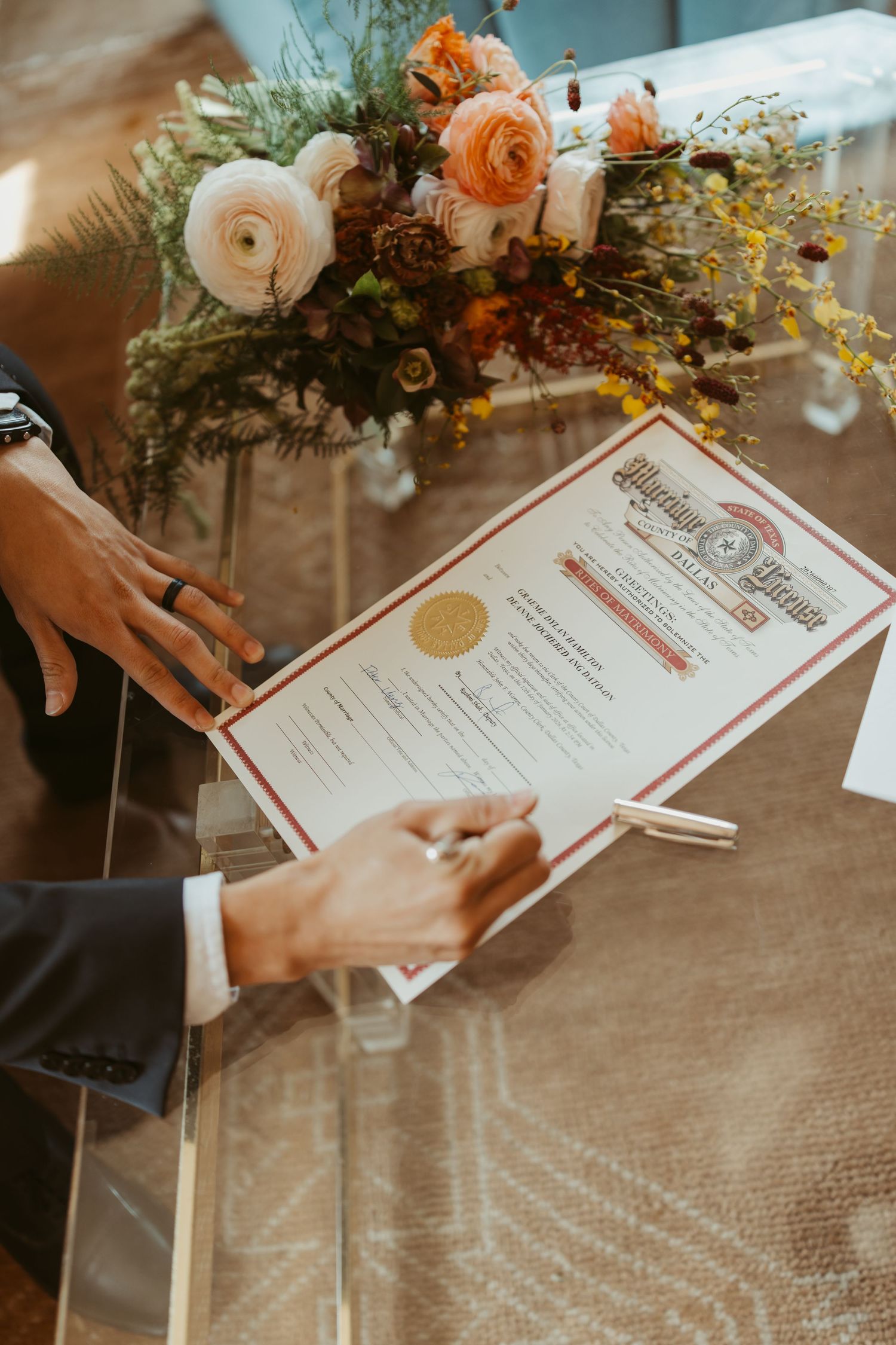 Person signing a marriage license certificate on a glass table with an elegant floral arrangement in the background.